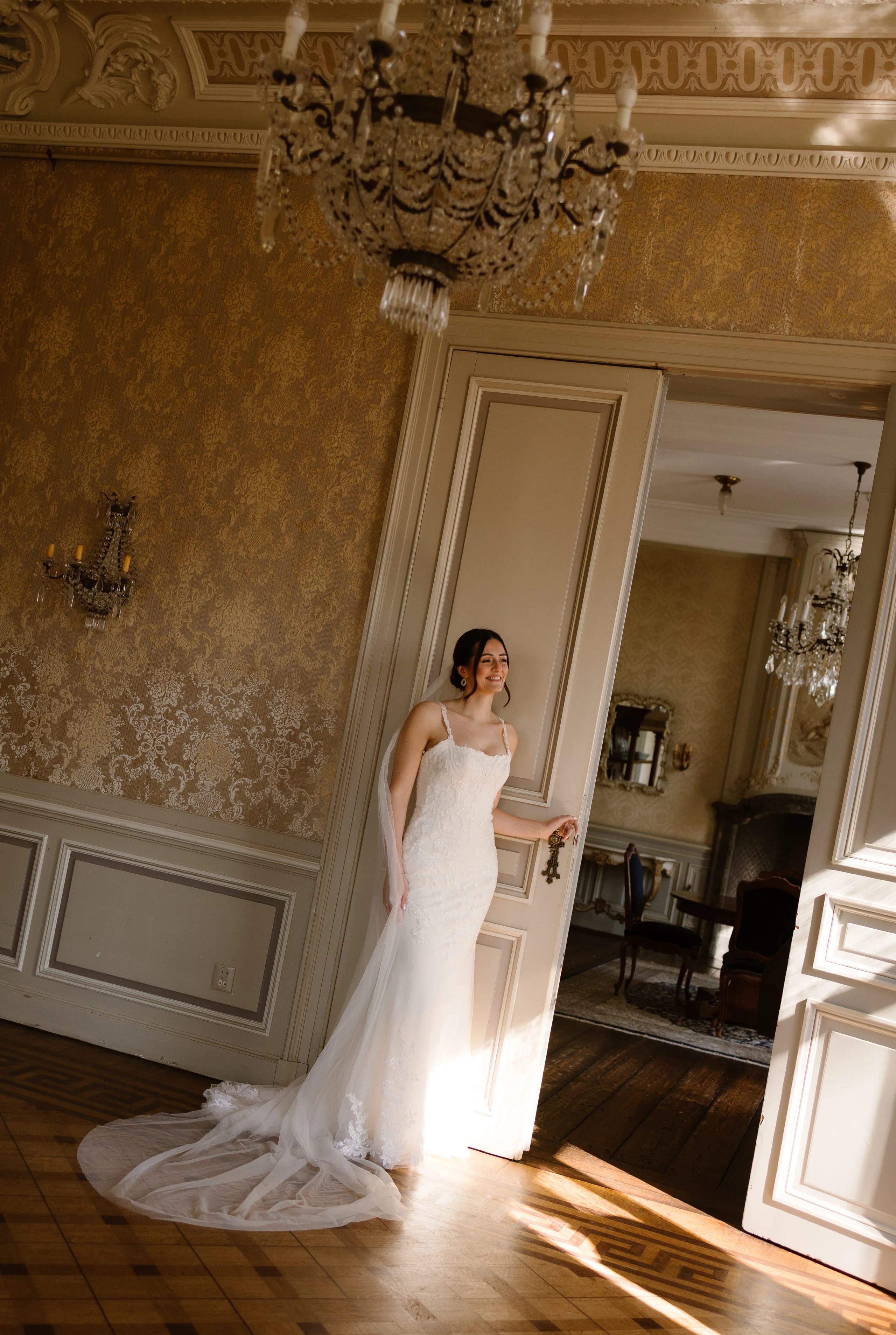 Een bruid in een elegante trouwjurk met een lange sleep, poseert bij een open deur in een sfeervolle, luxe kamer met gouden wanddecoraties en een kroonluchter.