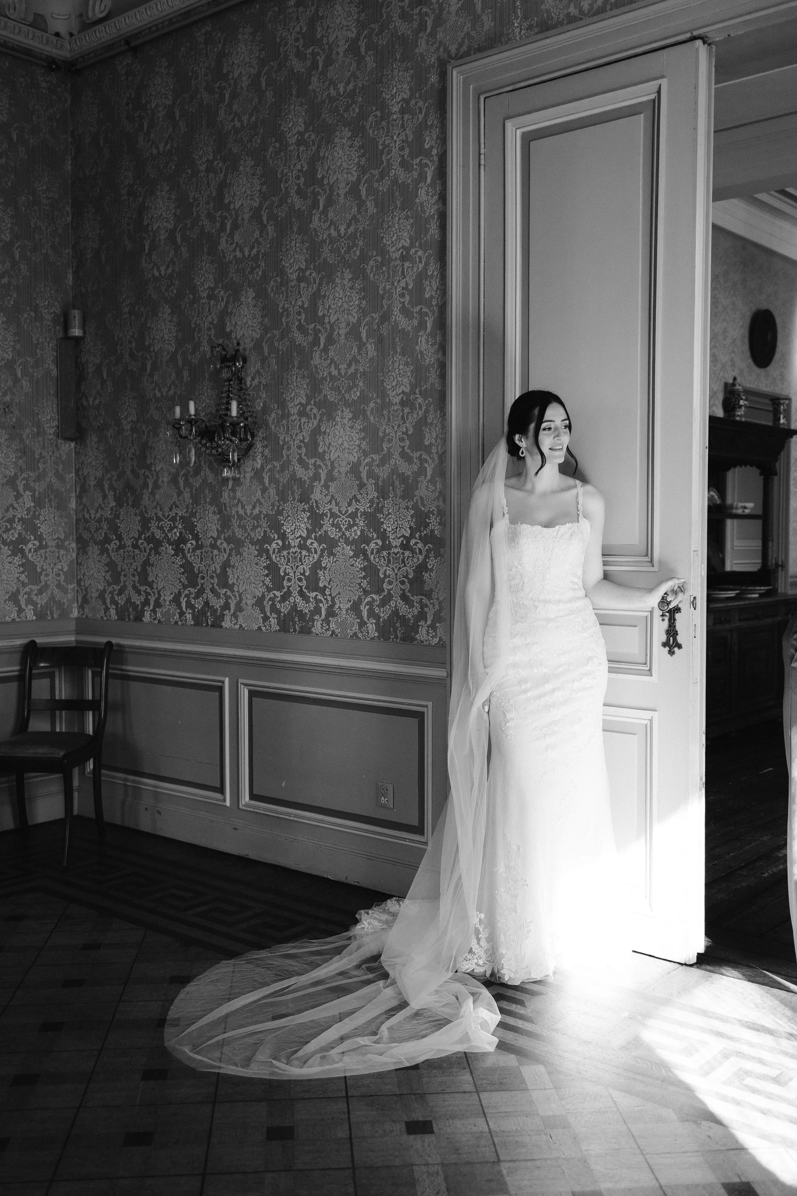 Een bruid in een elegant wit trouwkleed staat in de deuropening van een stijlvolle kamer, terwijl zonlicht subtie over de vloer valt en haar lange sluier accentueert. De nostalgische decoraties van de ruimte benadrukken de romantische sfeer van deze speciale gelegenheid.