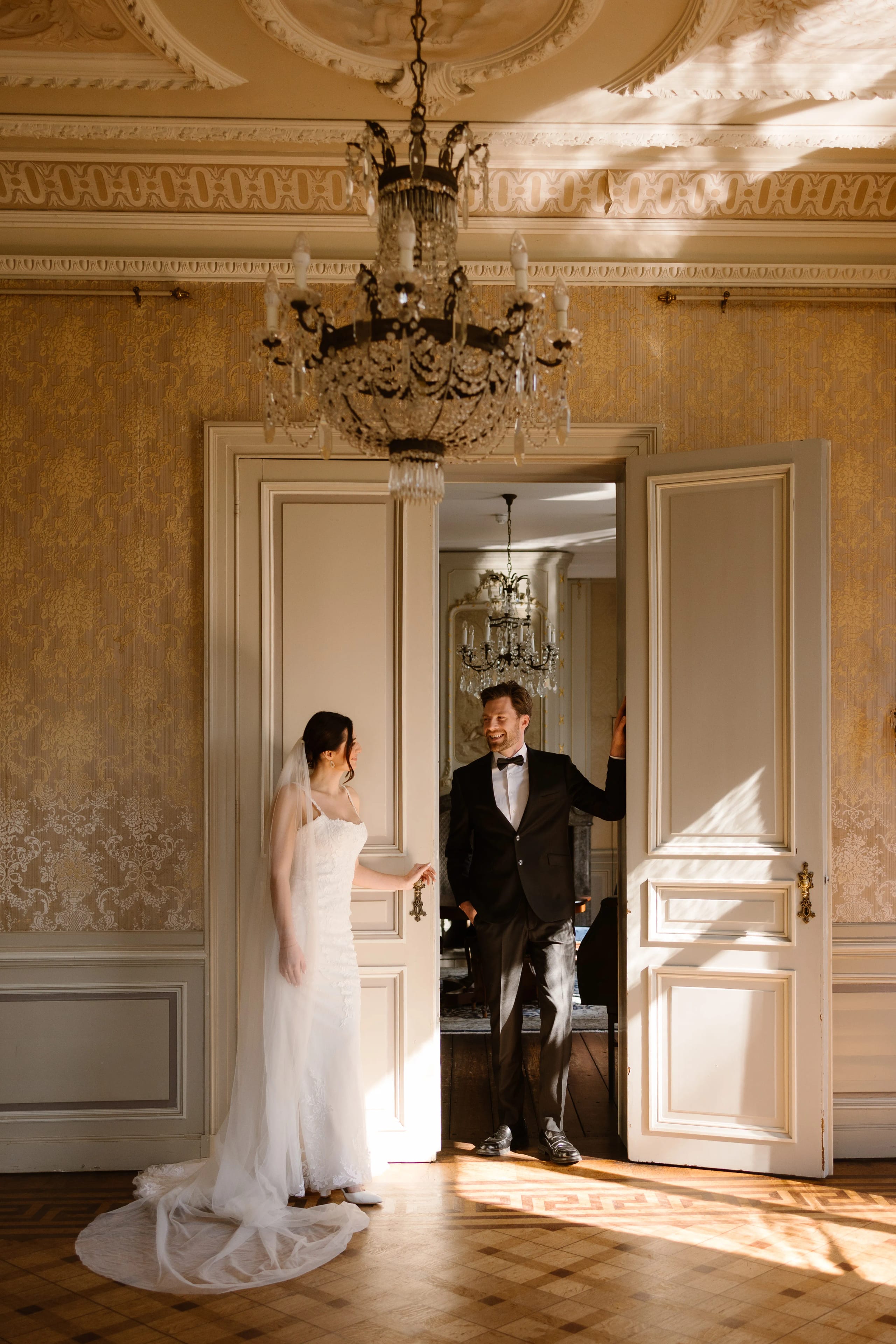 Een bruid in een elegante trouwjurk en een bruidegom in een zwart pak staan in een stijlvolle kamer met veel natuurlijke lichtinval, terwijl ze elkaar met een glimlach aankijken bij een open deur omringd door een chandelier en vergulde wanddecoraties.