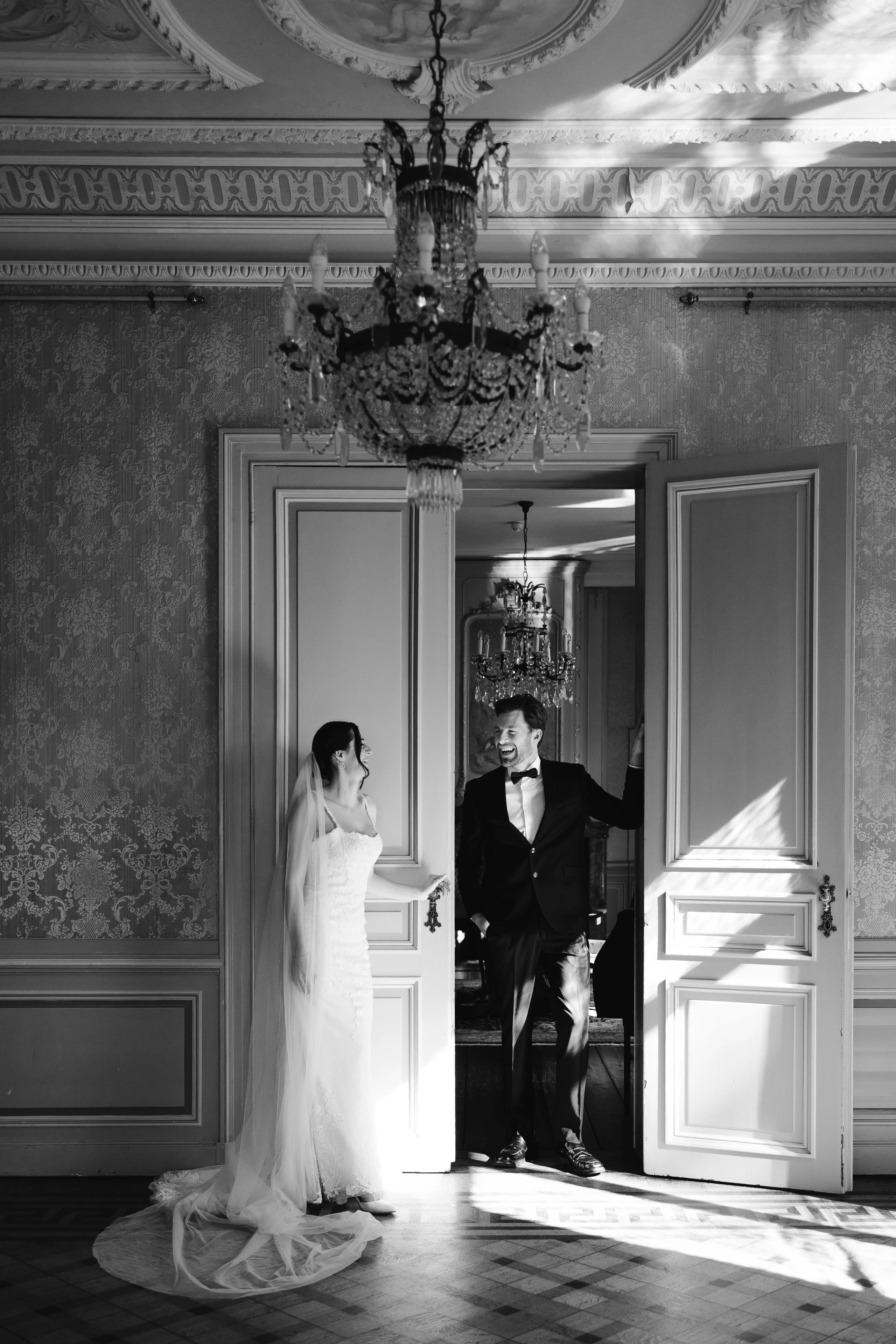 Een bruid in een elegante jurk met een sluier staat in een sfeervolle ruimte, terwijl de bruidegom in een zwart kostuum met een strikje leunt tegen een open deur. Het zachte licht valt naar binnen en creëert een romantische ambiance.