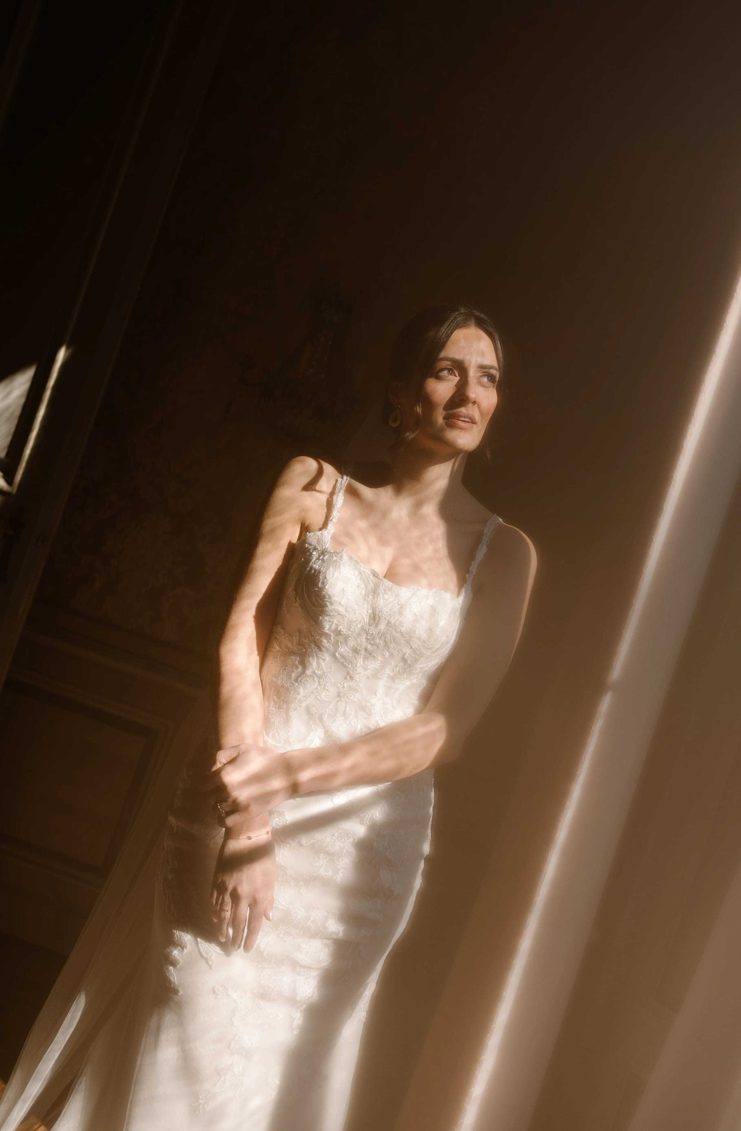 Een elegante bruid in een prachtig kant-en-bloemend trouwjurk poseert in zacht, warm licht dat door een raam valt, met een serene uitstraling en sierlijke accessoires.