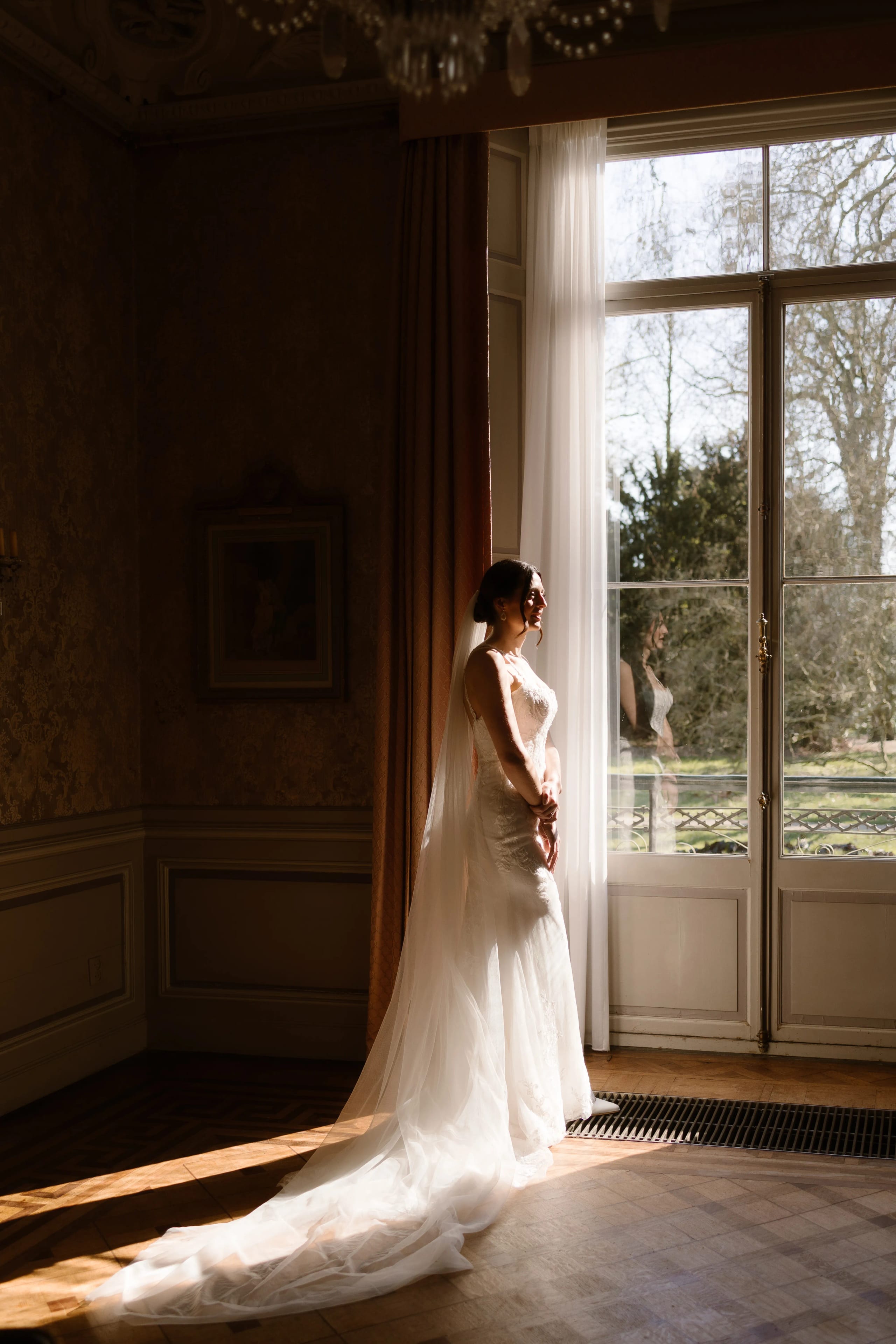 Een bruid in een elegante trouwjurk staat bij een grote raam, terwijl natuurlijk licht naar binnen valt en schaduw op de vloer werpt. De serene sfeer wordt versterkt door de prachtige achtergrond met uitzicht op een groene tuin.