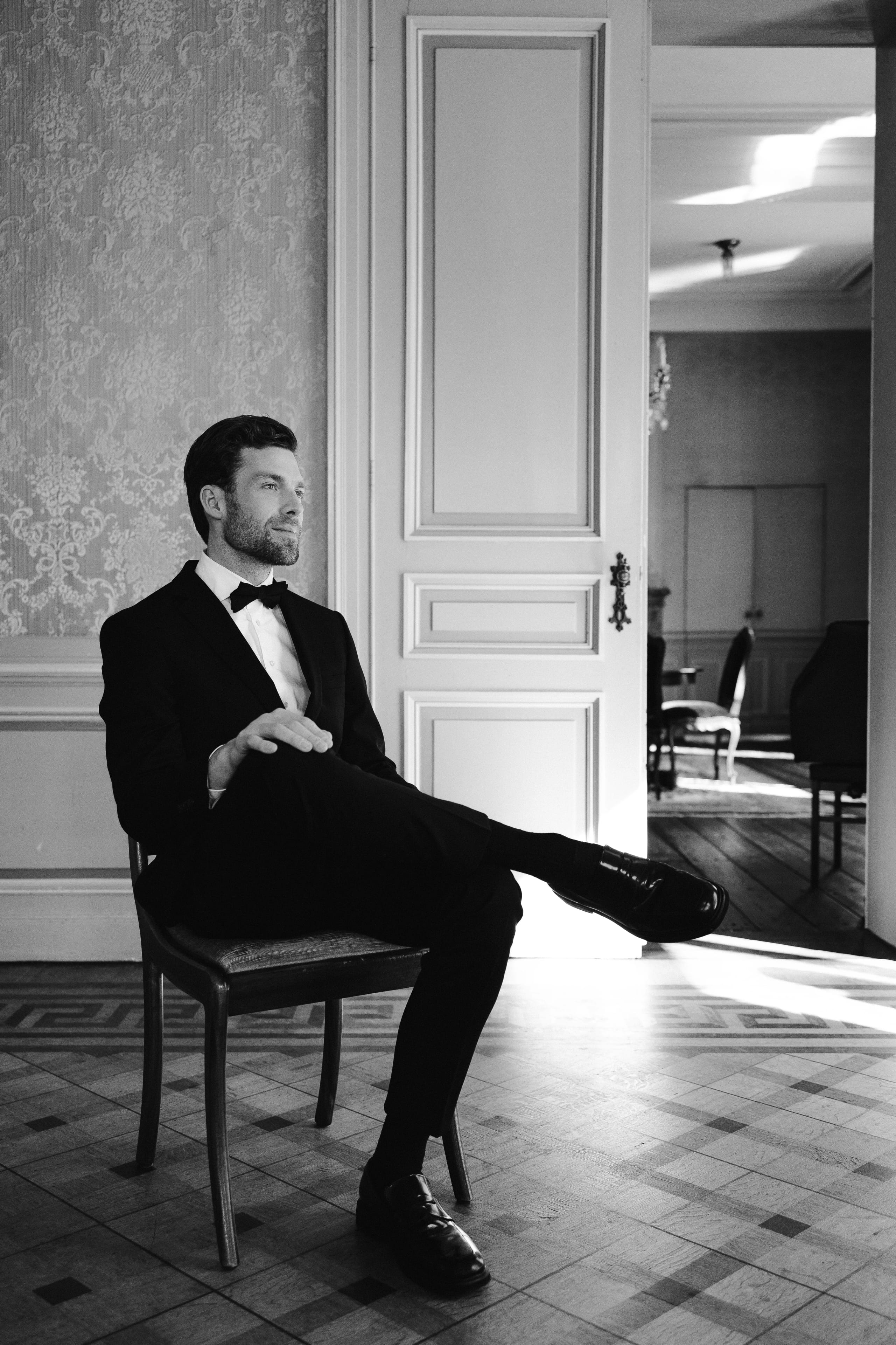 Een stijlvol geklede man in smoking en strikje zit ontspannen op een stoel, met zijn benen over elkaar geslagen, terwijl hij in een elegant ingerichte ruimte met veel lichtinval kijkt.