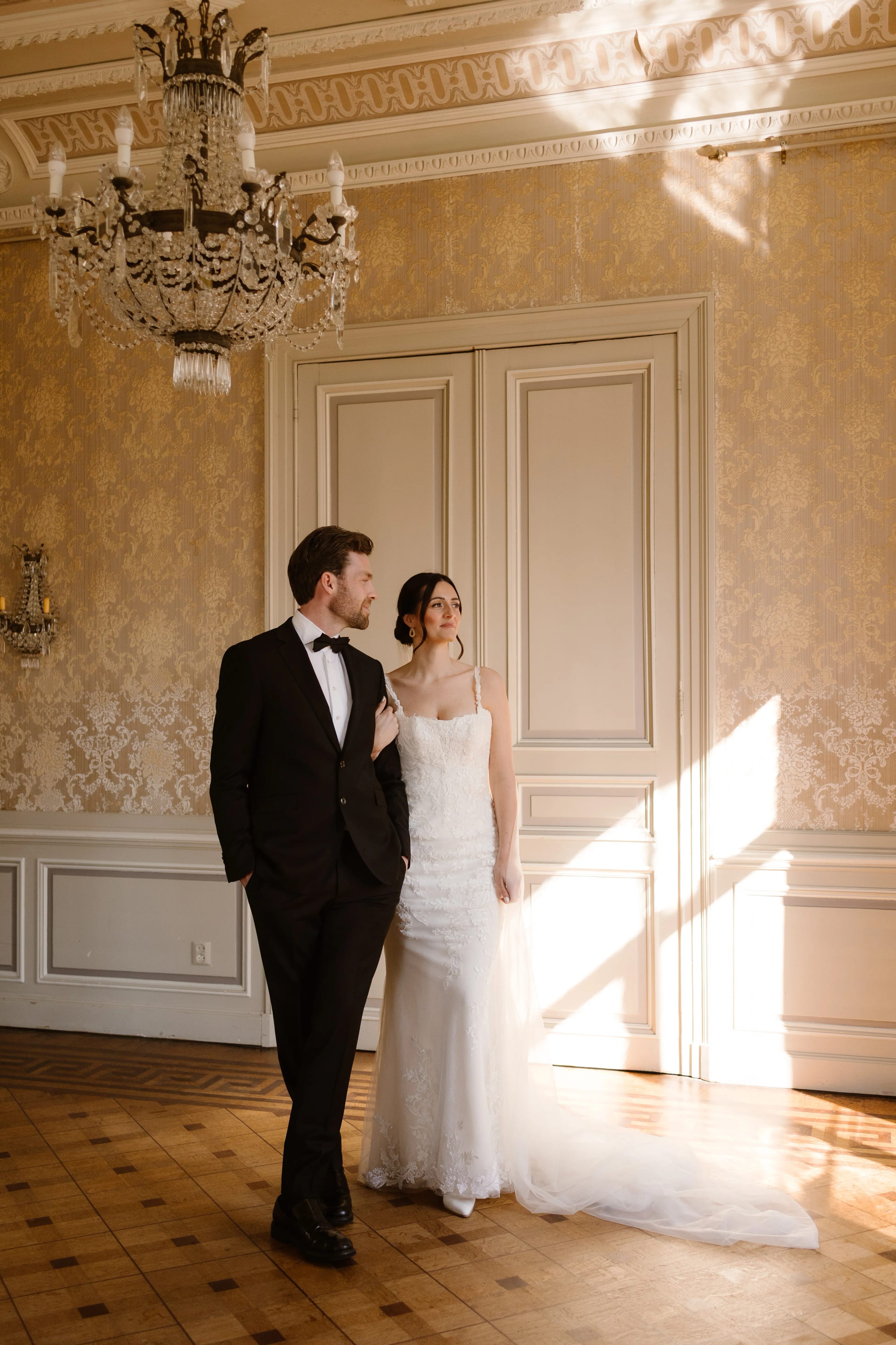 Een elegant bruidspaar poseert in een prachtig aangeklede ruimte, met een chique kroonluchter boven hen en zacht natuurlijk licht dat door het raam valt, waardoor de romantische sfeer van hun speciale dag wordt benadrukt.
