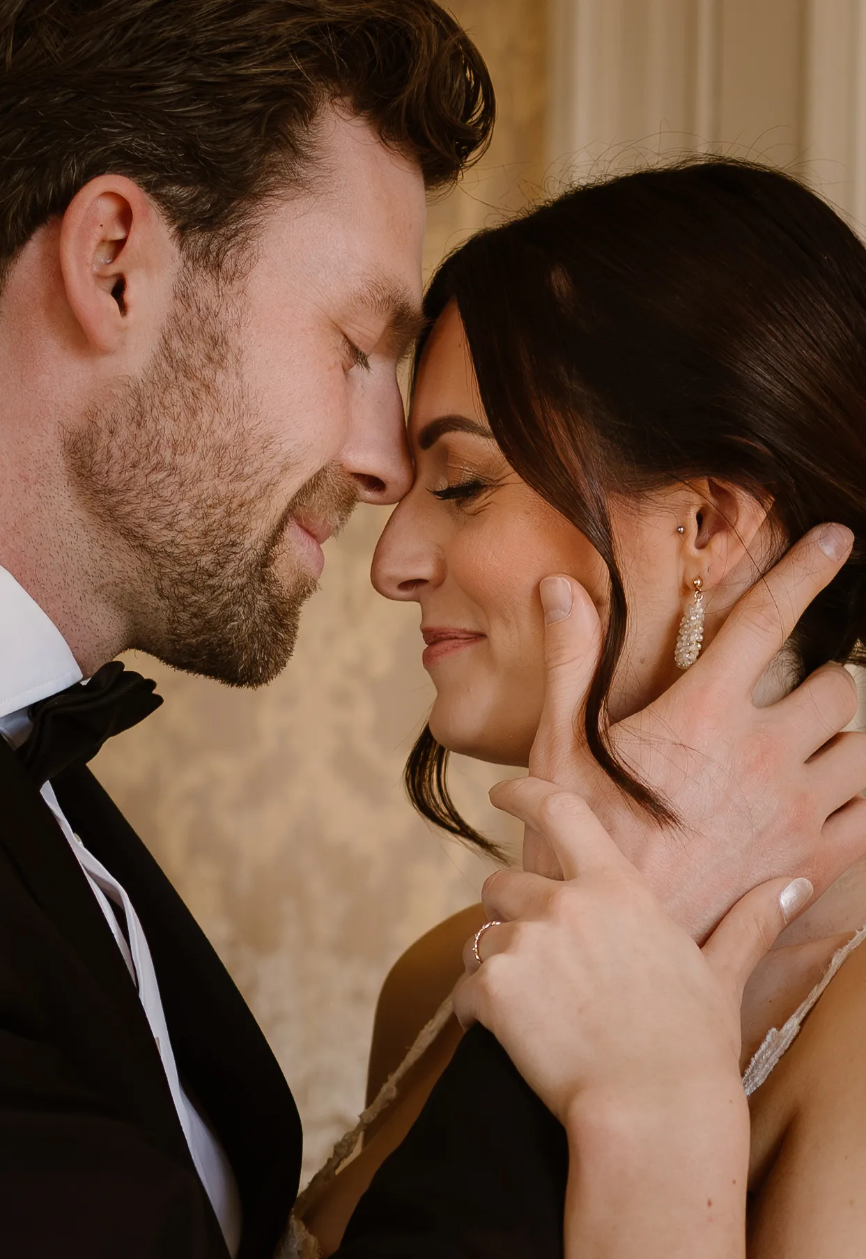 Een romantisch stel geniet van een intiem moment terwijl ze liefdevol naar elkaar kijken en elkaars gezichten aanraken, gekleed in formele kleding tegen een elegante achtergrond.