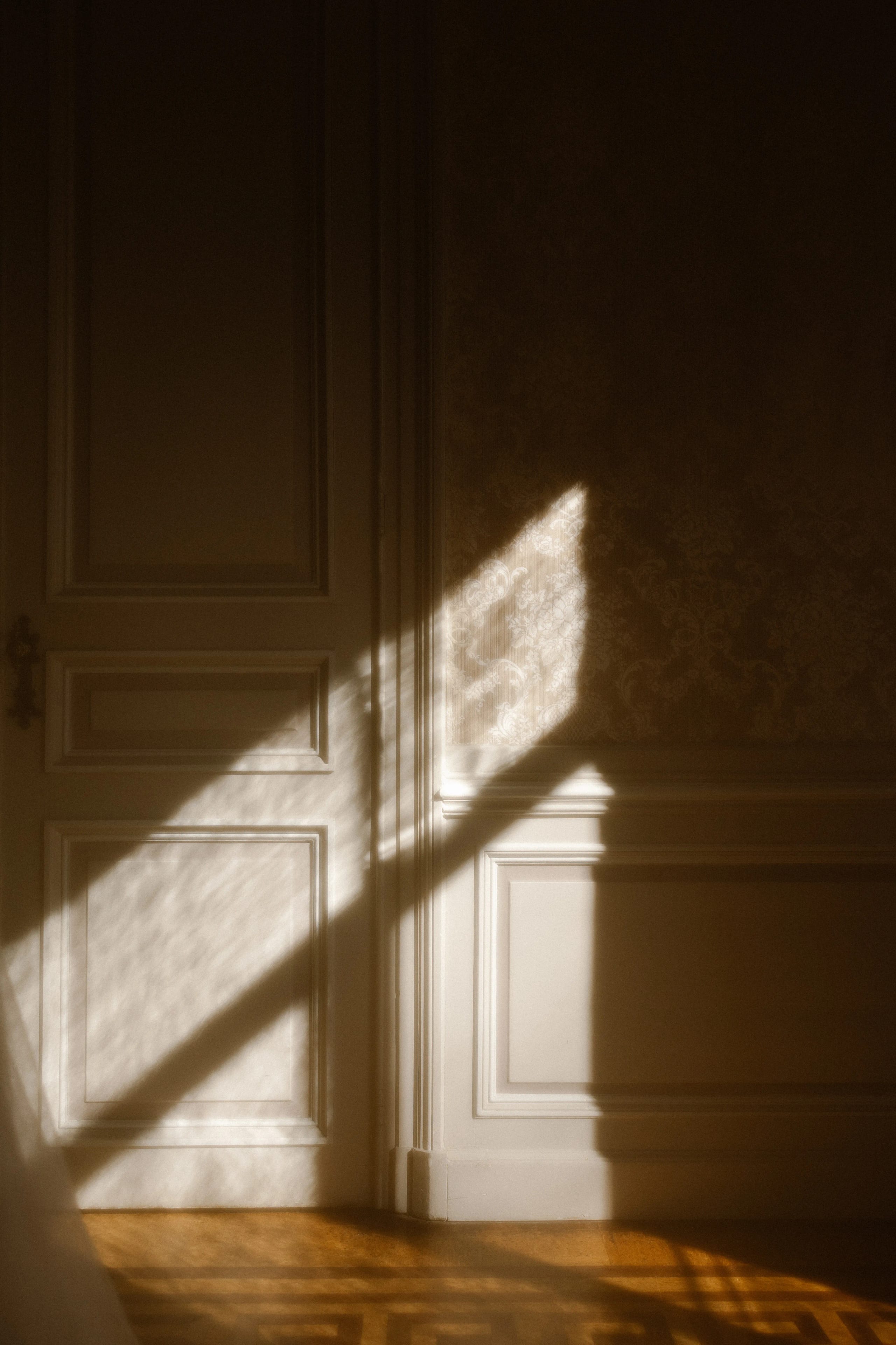 Zacht zonlicht valt door een deur op een muur, waardoor een speelse schaduw en een warme sfeer in de kamer ontstaat, met een uitstraling van elegantie en rust.