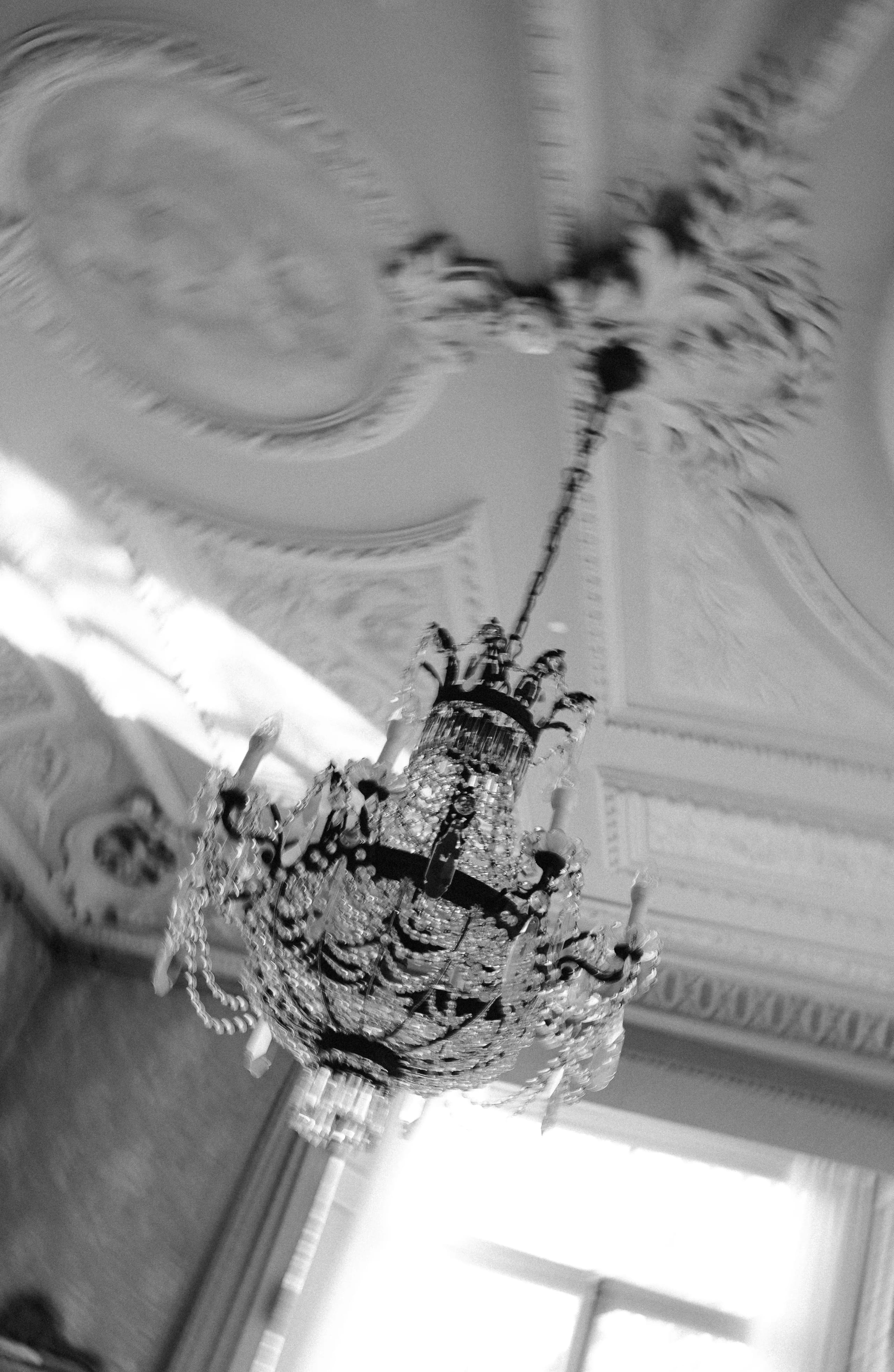 Een elegante kristallen kroonluchter hangt aan het plafond van een weelderige kamer, met gedetailleerde ornamenten en mooi licht dat door het raam naar binnen valt.