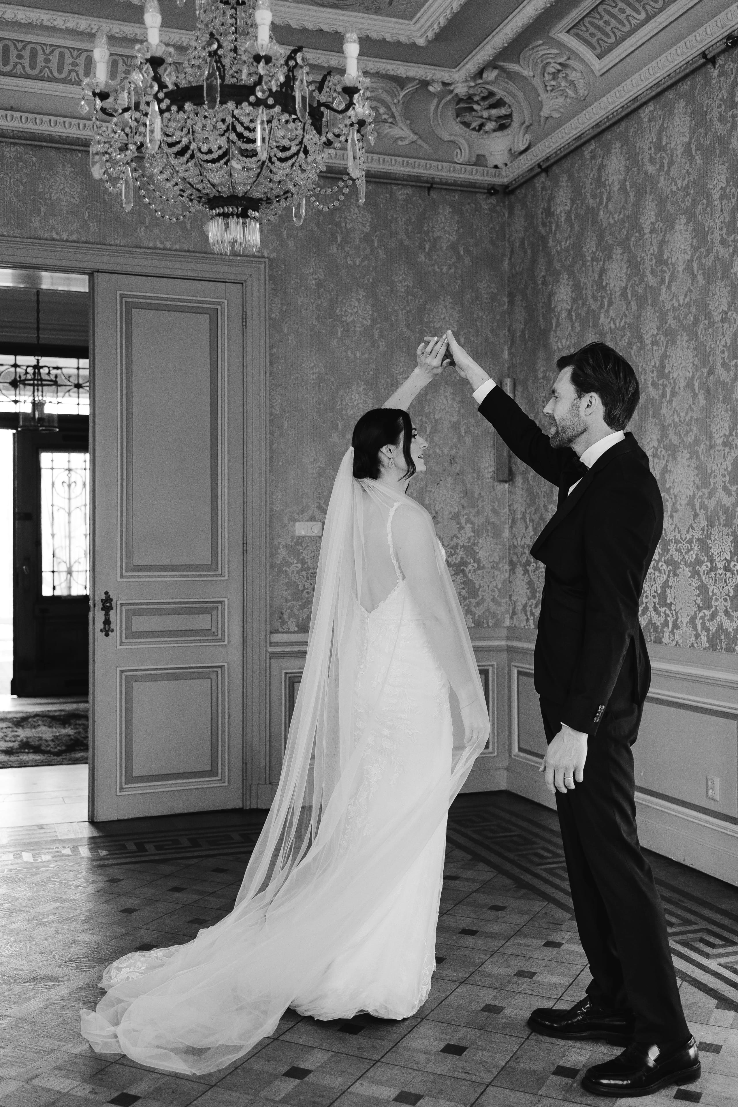 Een gelukkige bruid en bruidegom dansen samen in een elegant ingerichte ruimte, terwijl de bruidegom haar hand omhoog houdt in een romantische vlucht. De luxe omgeving met een kroonluchter en gedetailleerde wandversieringen voegt een vleugje grandeur toe aan hun speciale moment.