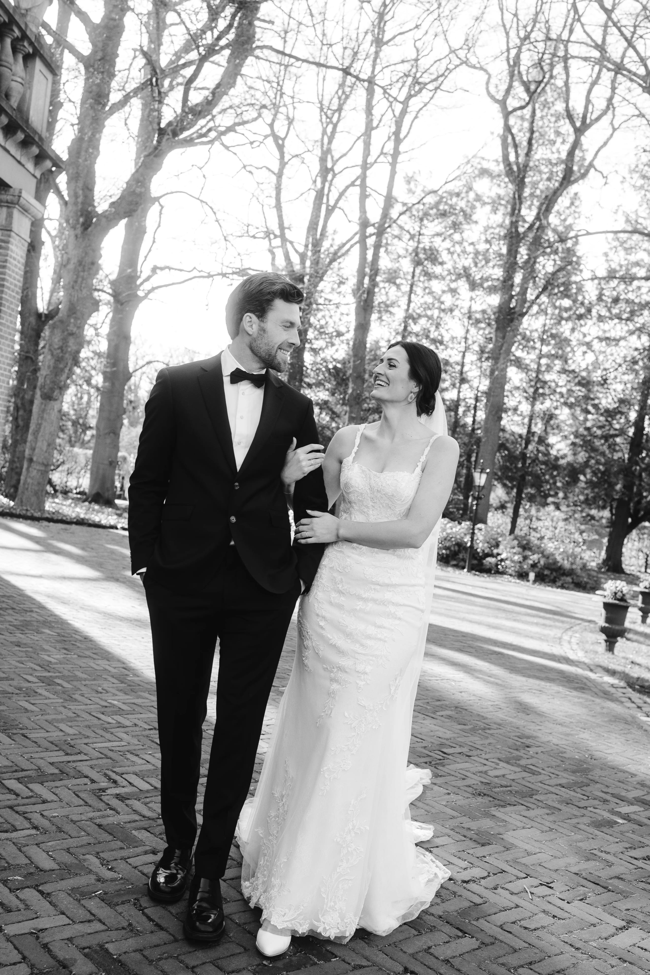 Een gelukkig bruidspaar wandelt hand in hand door een mooi park, omringd door bomen, terwijl ze naar elkaar lachen. De bruid draagt een elegante, witte trouwjurk en de bruidegom een stijlvol zwart pak met een strik.