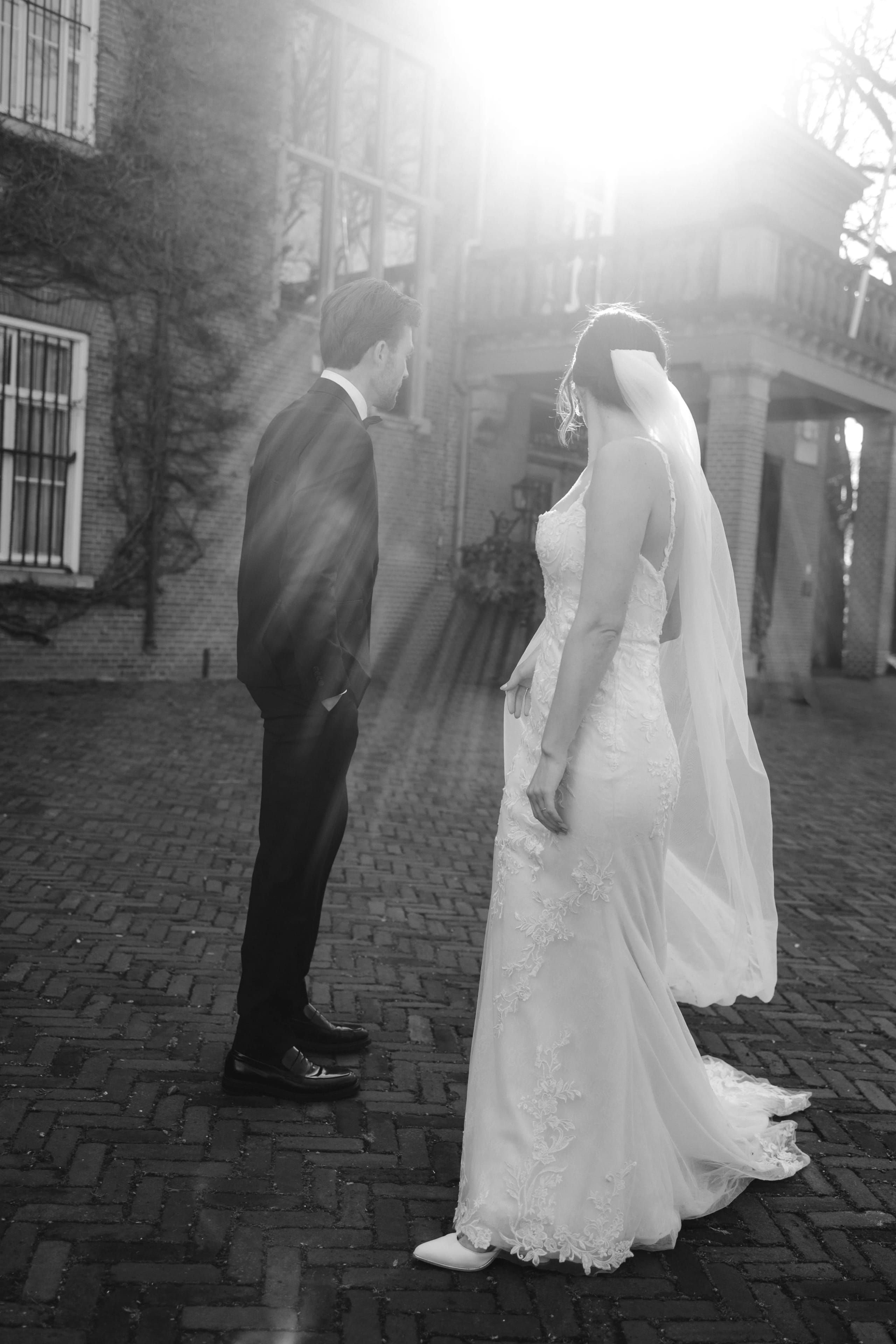 Een bruid in een schitterende, witte trouwjurk en met een sluier, die aan het eind van een sierlijke straat staat, kijkt naar haar aanstaande echtgenoot in een zwart pak, terwijl de zon fel achter hen schijnt en het romantische moment versterkt.