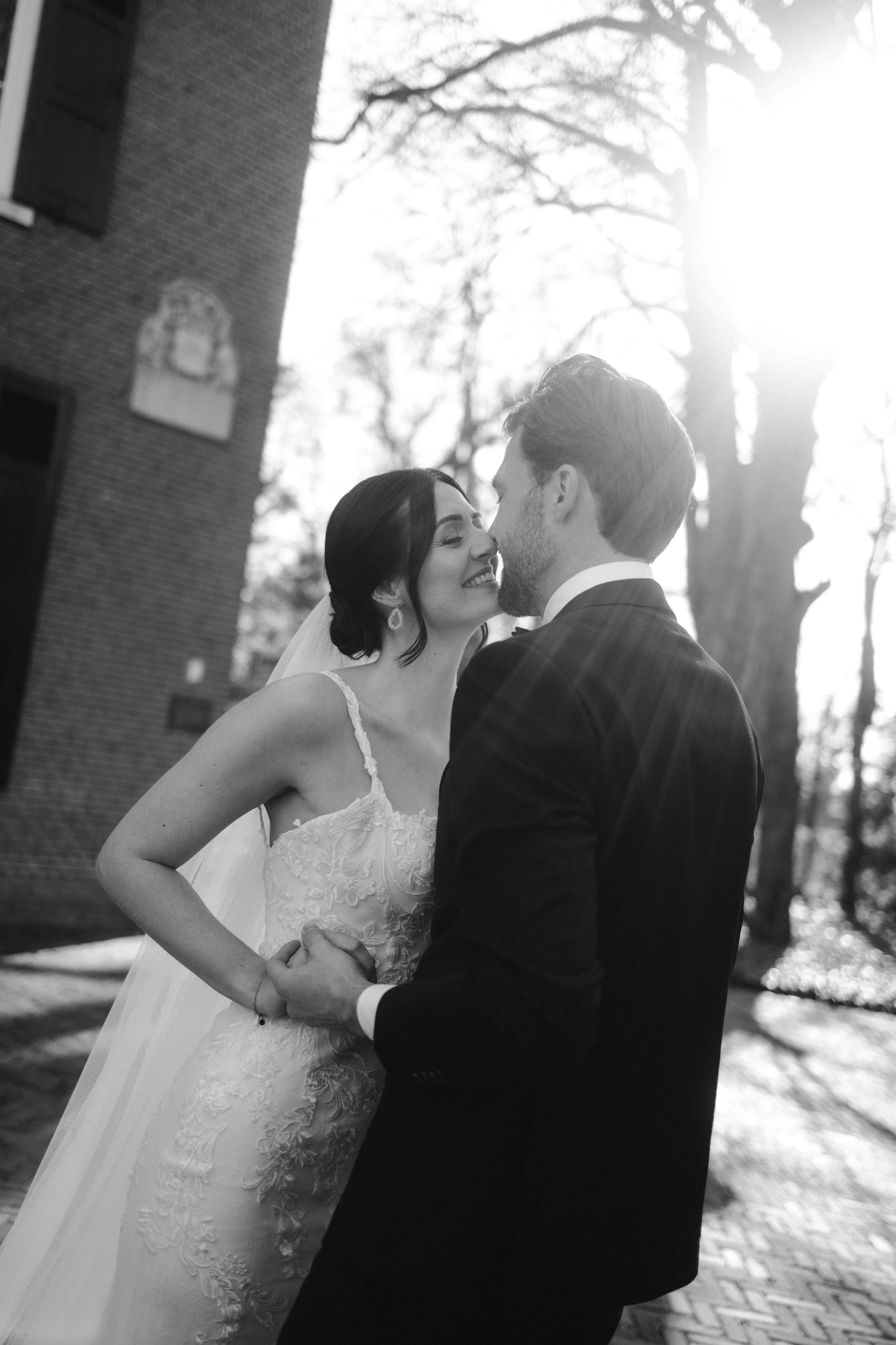 Een gelukkig bruidspaar deelt een intieme kus tijdens hun huwelijksceremonie, omringd door een romantische buitenomgeving met zachte zonnestralen die door de bomen schijnen.