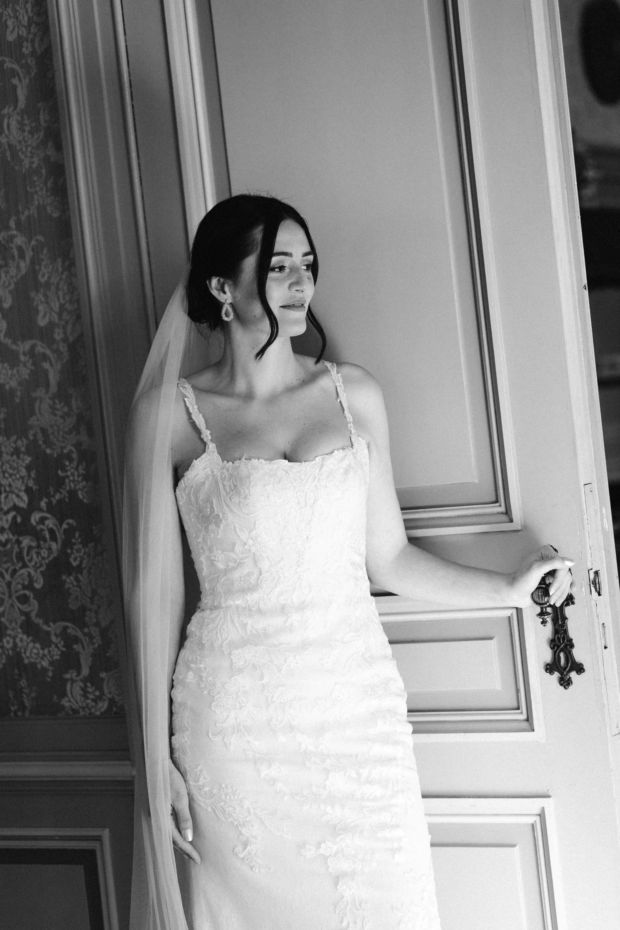 Een elegante bruid in een prachtig kanten trouwjurk staat bij een geopend deurtje, met een sluier die soepel over haar schouders valt terwijl ze vol vertrouwen kijkt.