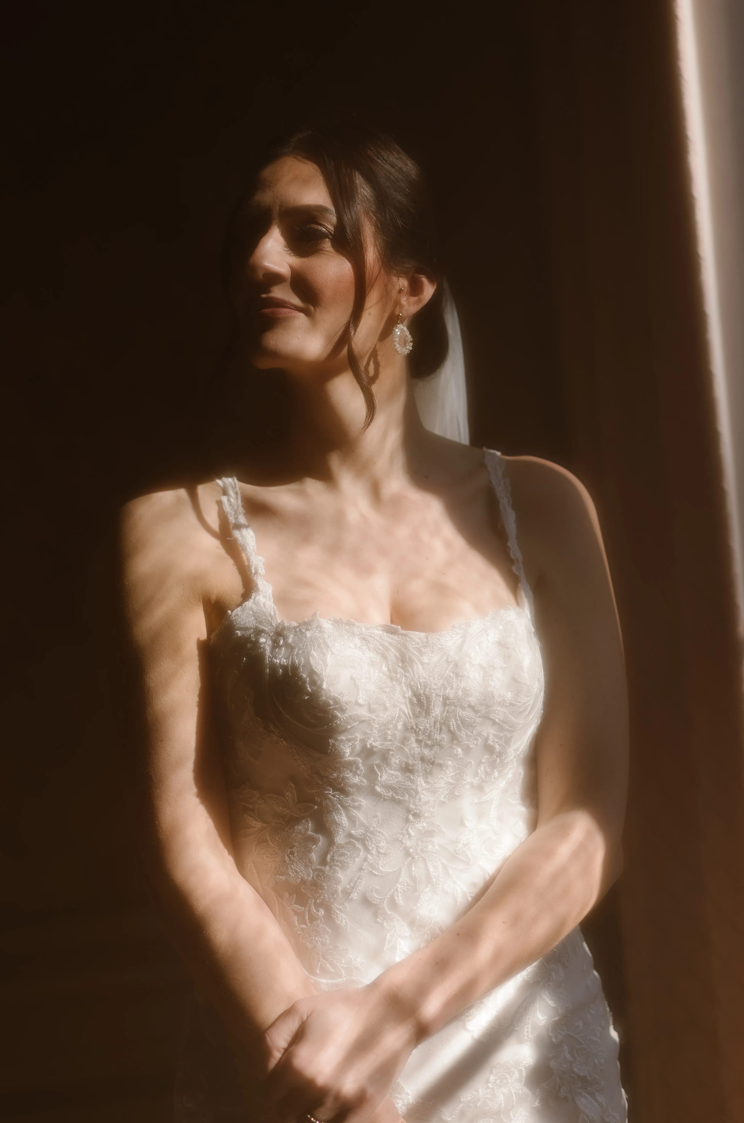 Een prachtige bruid in een schitterende, met pailletten versierde trouwjurk poseert sfeervol voor een raam, terwijl het zachte licht een romantische glans op haar gezicht werpt.