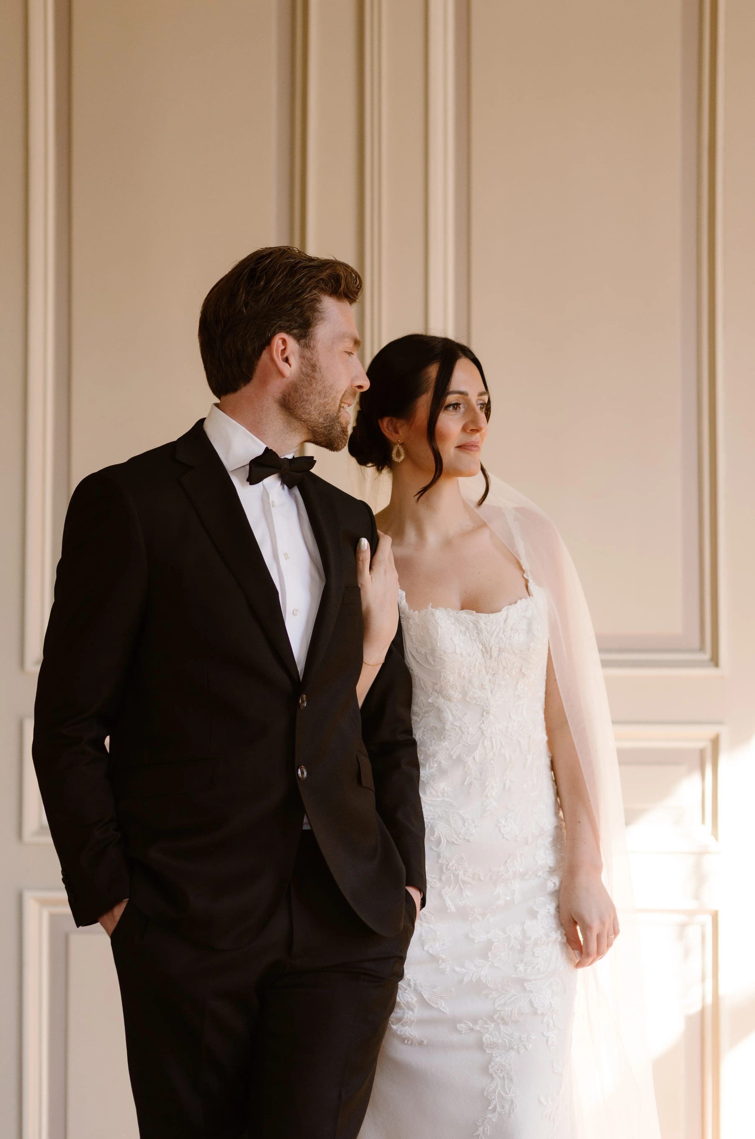 Een bruidsstel poseert elegant voor een lichte achtergrond, met de bruid in een prachtig, gedetailleerd trouwjurk og de bruidegom gekleed in een klassieke zwart kostuum met een strik.