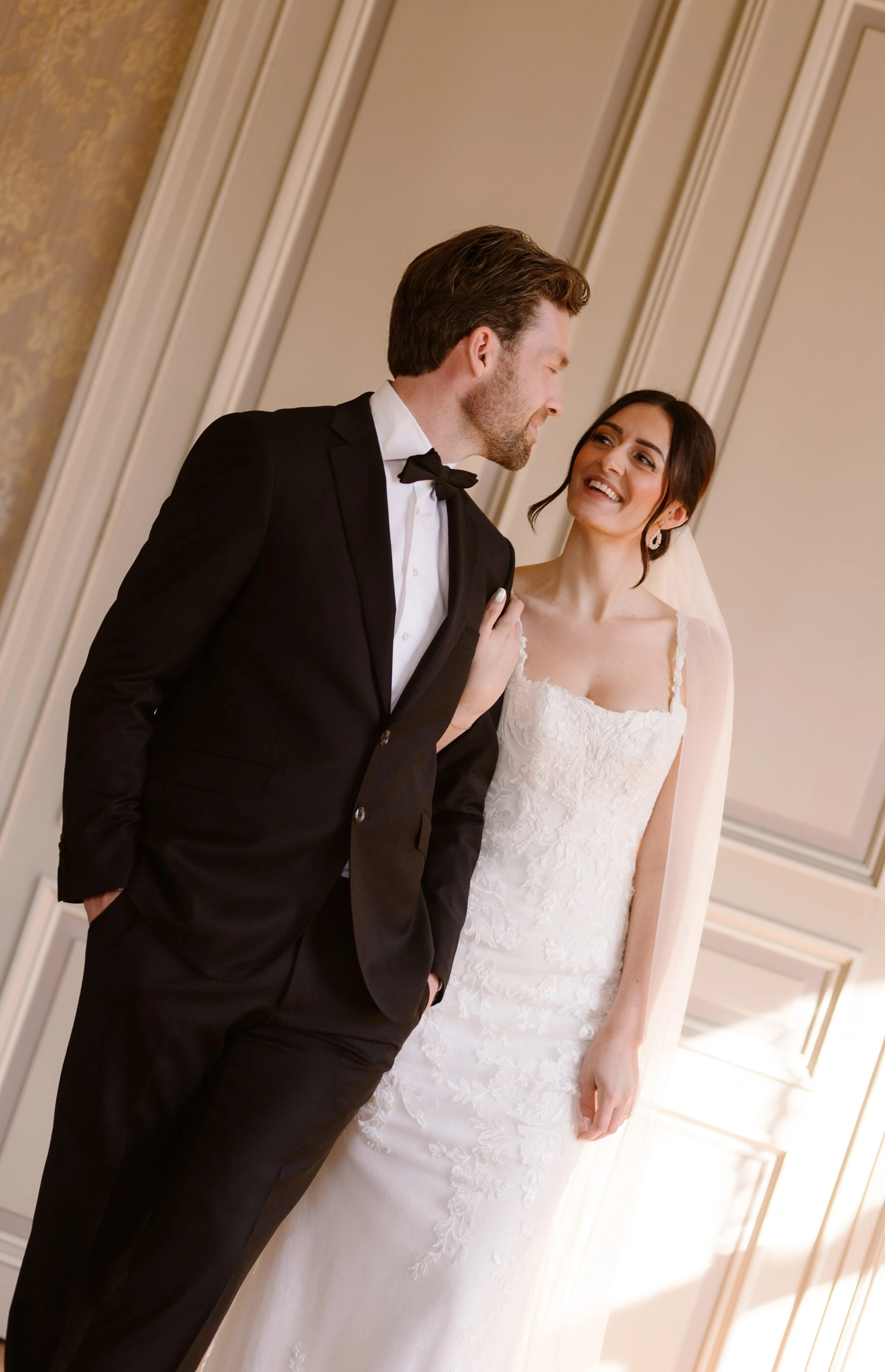 Een gelukkig bruidspaar poseert in een elegante setting, waarbij de bruid in een fraaie witte trouwjurk met kant staat en de bruidegom in een klassieke zwarte tuxedo. Hun blikken spreken liefde en vreugde uit, met warme glimlachen terwijl ze dicht bij elkaar staan.