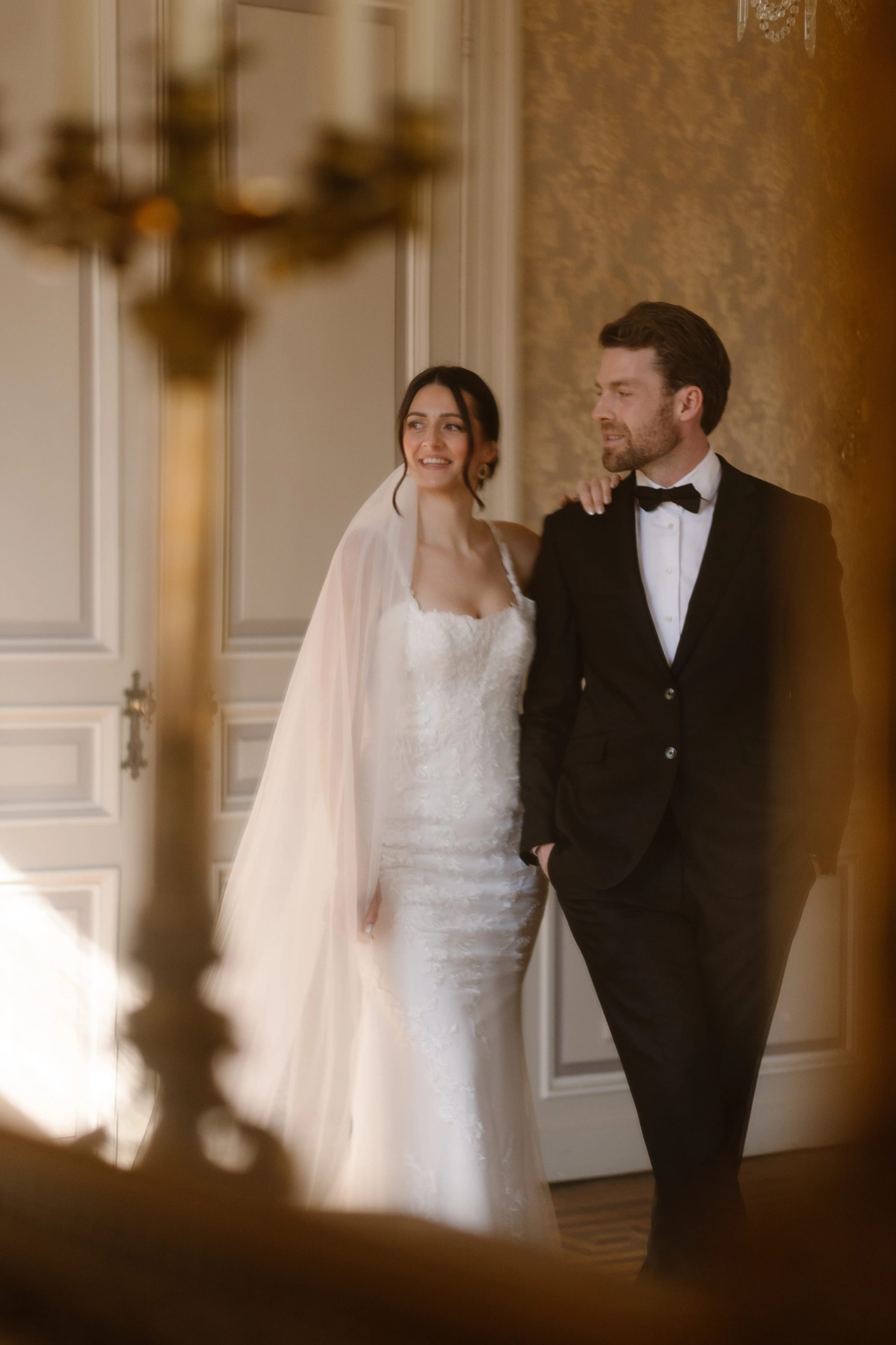 Een bruid in een elegante trouwjurk en een bruidegom in een zwart pak lopen samen, met liefdevolle blikken naar elkaar. De achtergrond is een prachtig versierd interieur met warme verlichting, wat de romantische sfeer van het moment versterkt.