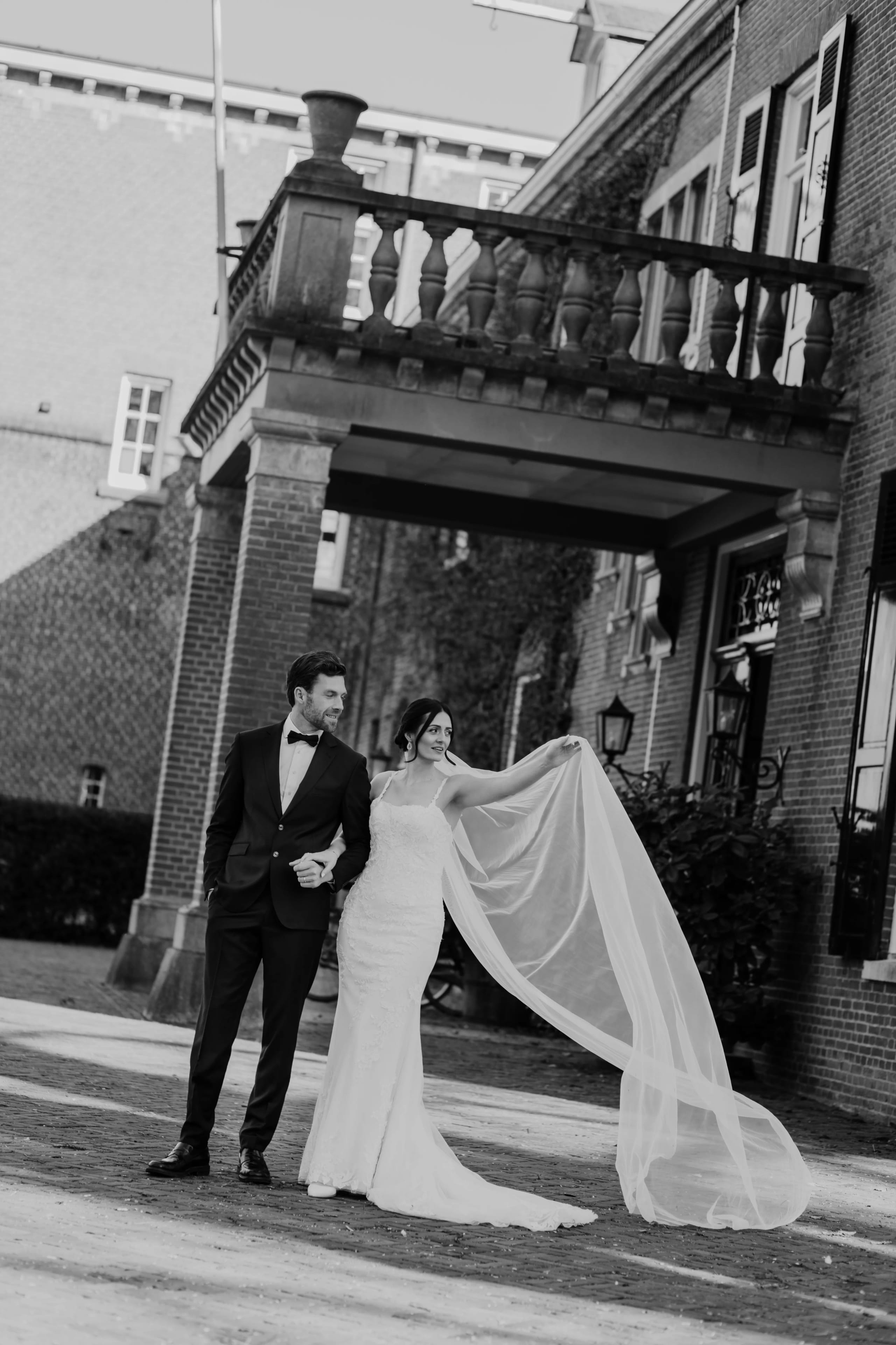 Een bruid en bruidegom staan hand in hand voor een elegant gebouw, terwijl de bruid speels haar lange sluier omhoog houdt, wat een romantische en feestelijke sfeer creëert.