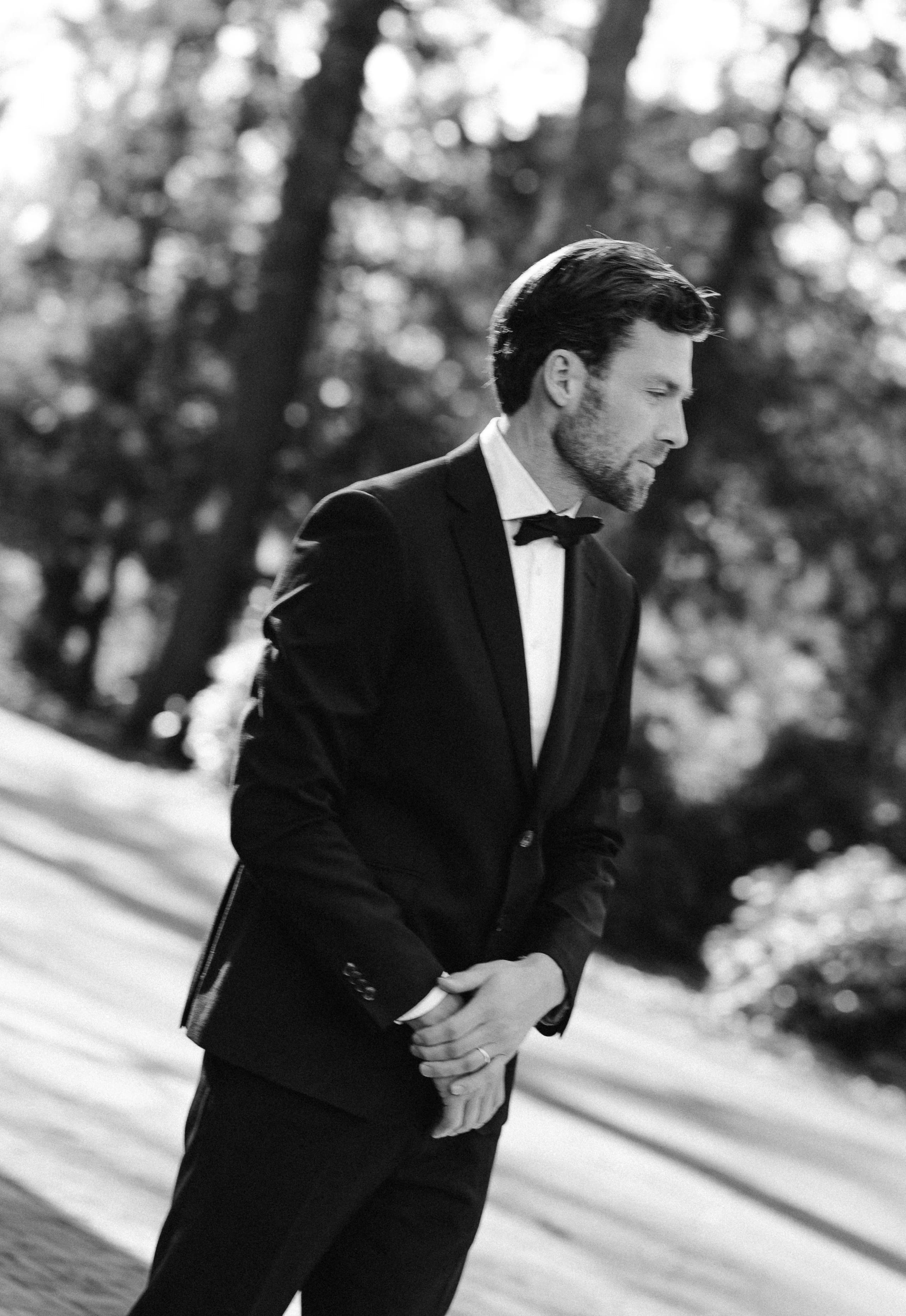 Een stijlvolle man in een zwart pak met een strikje, staande in een natuurlijke omgeving, omringd door bomen en greenery, met een contemplatieve uitdrukking.