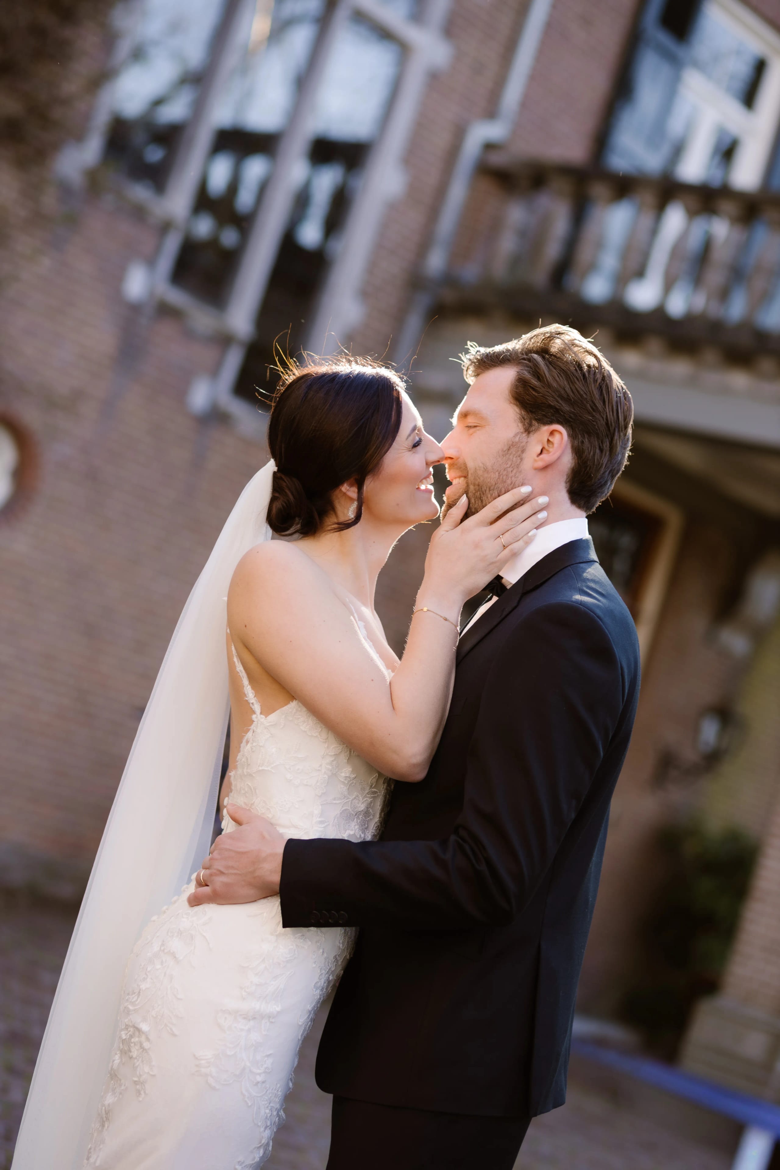 Een gelukkig bruidspaar kust elkaar liefdevol in de buitenlucht, omgeven door een romantische sfeer met een prachtig huis op de achtergrond. De bruid draagt een verfijnd, gedetailleerd trouwkleed, terwijl de bruidegom een elegante zwarte tuxedo aan heeft.
