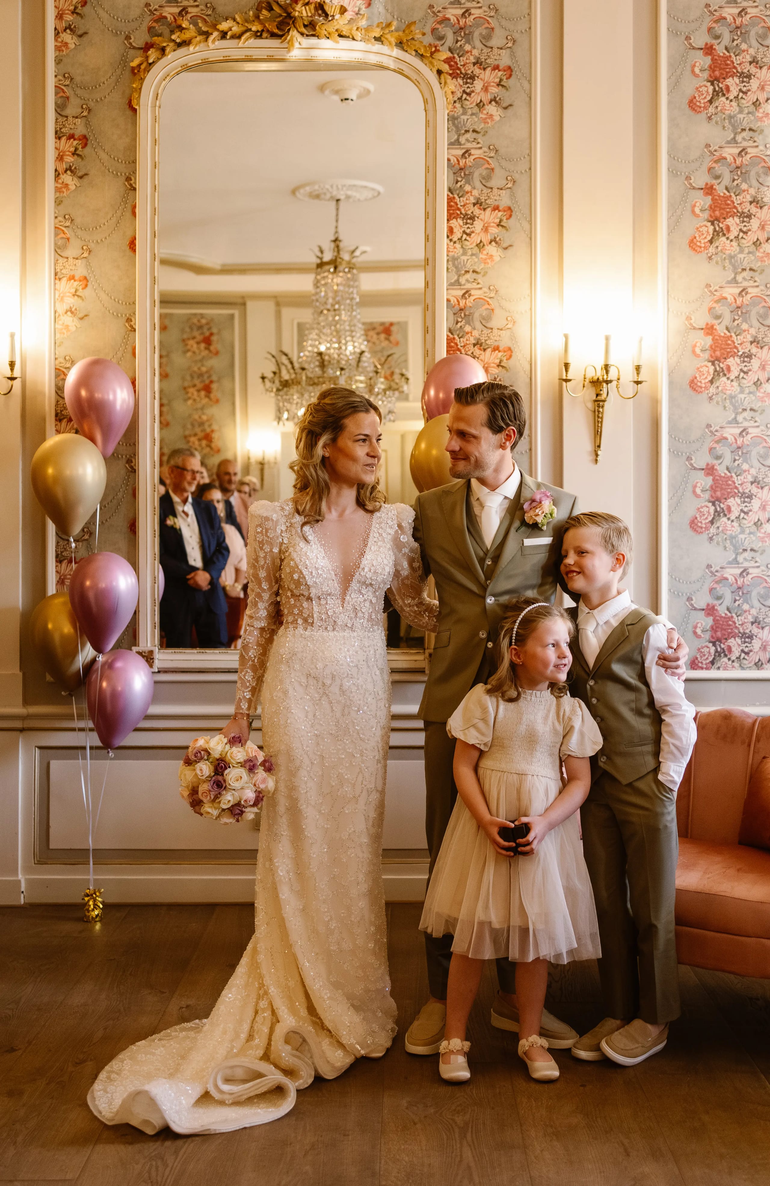 Bruid, bruidegom en twee kinderen poseren gelukkig voor een grote antieke spiegel