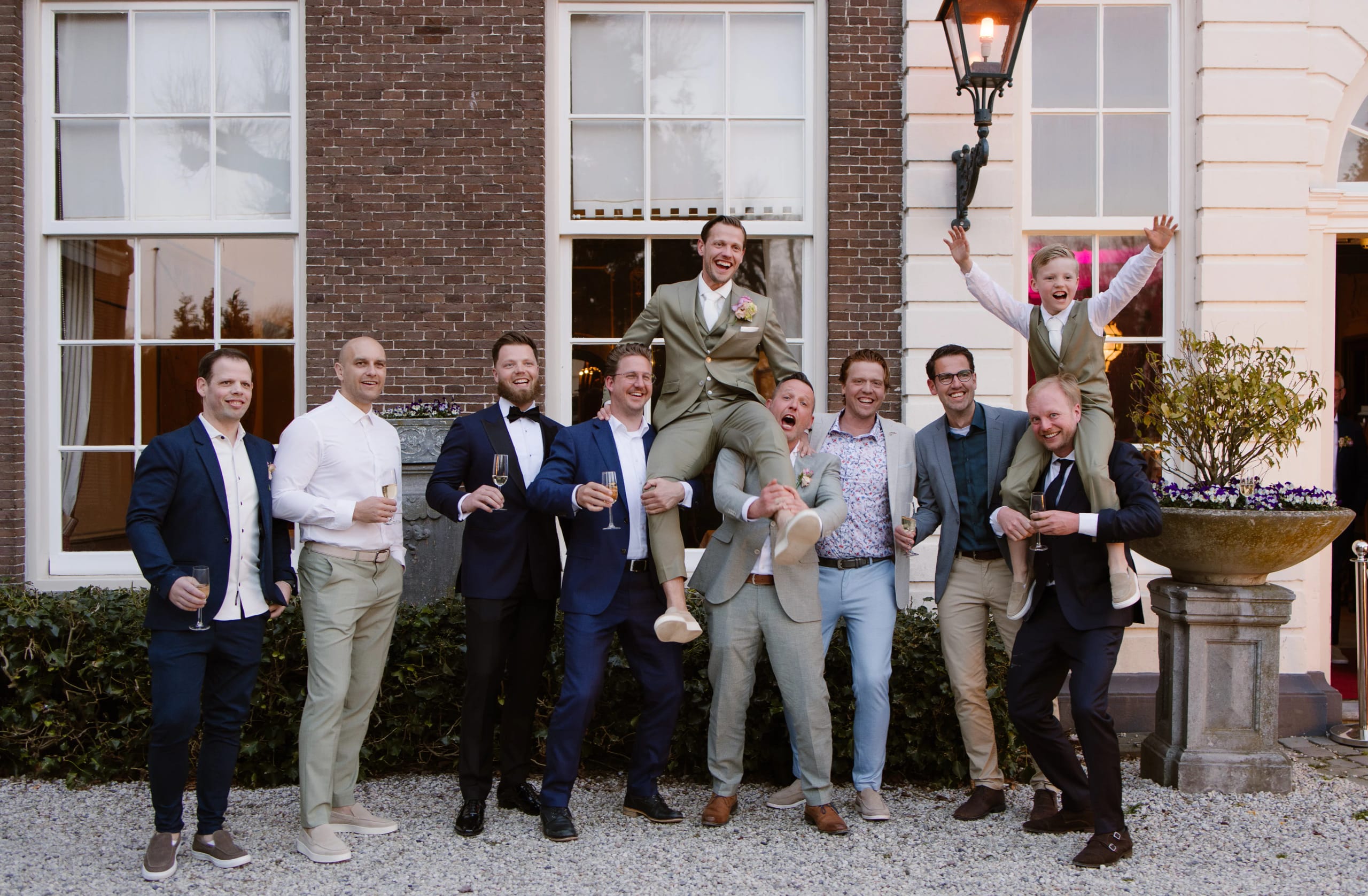 Lachende bruidegom in saliegroen pak wordt buiten in de lucht getild door vrienden