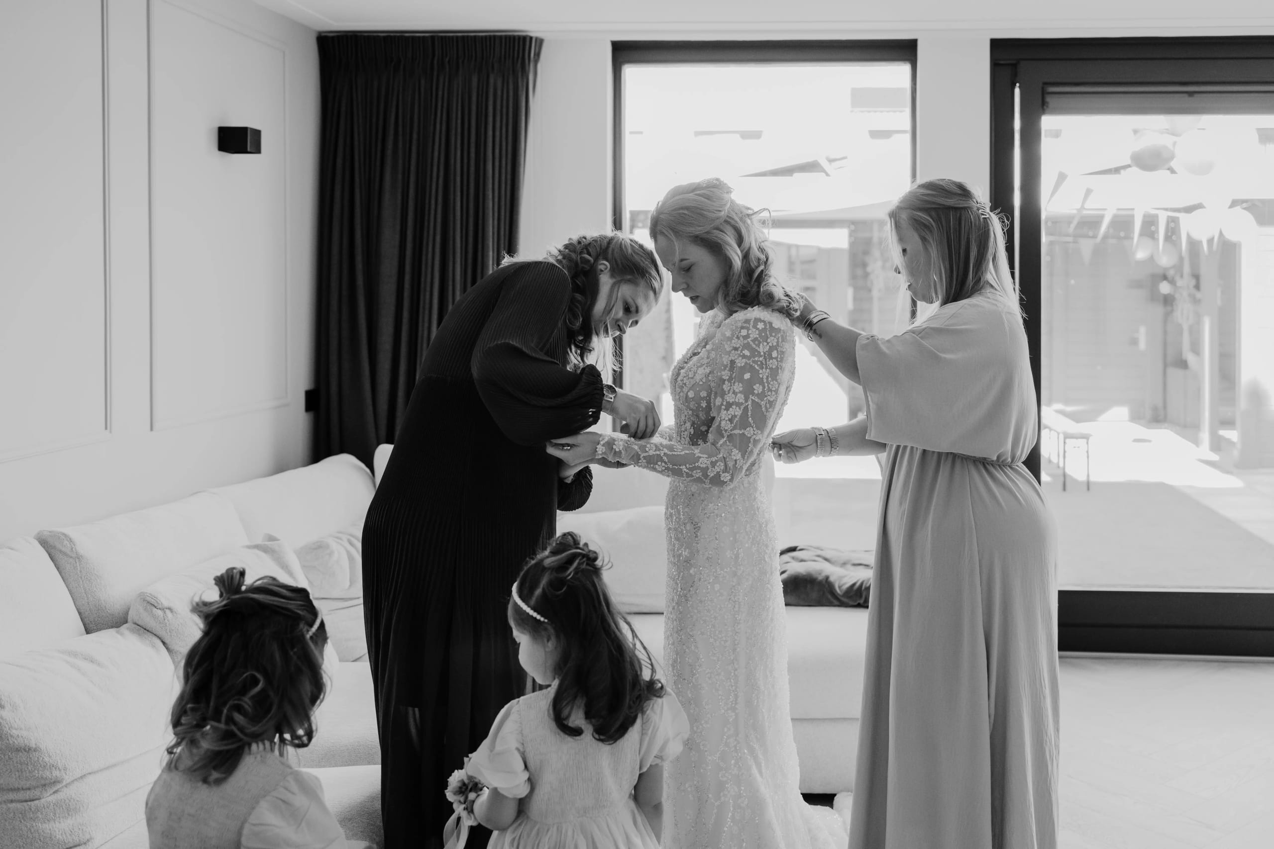 Zwart-wit foto van de bruid die geholpen wordt met het aantrekken van haar glinsterende jurk