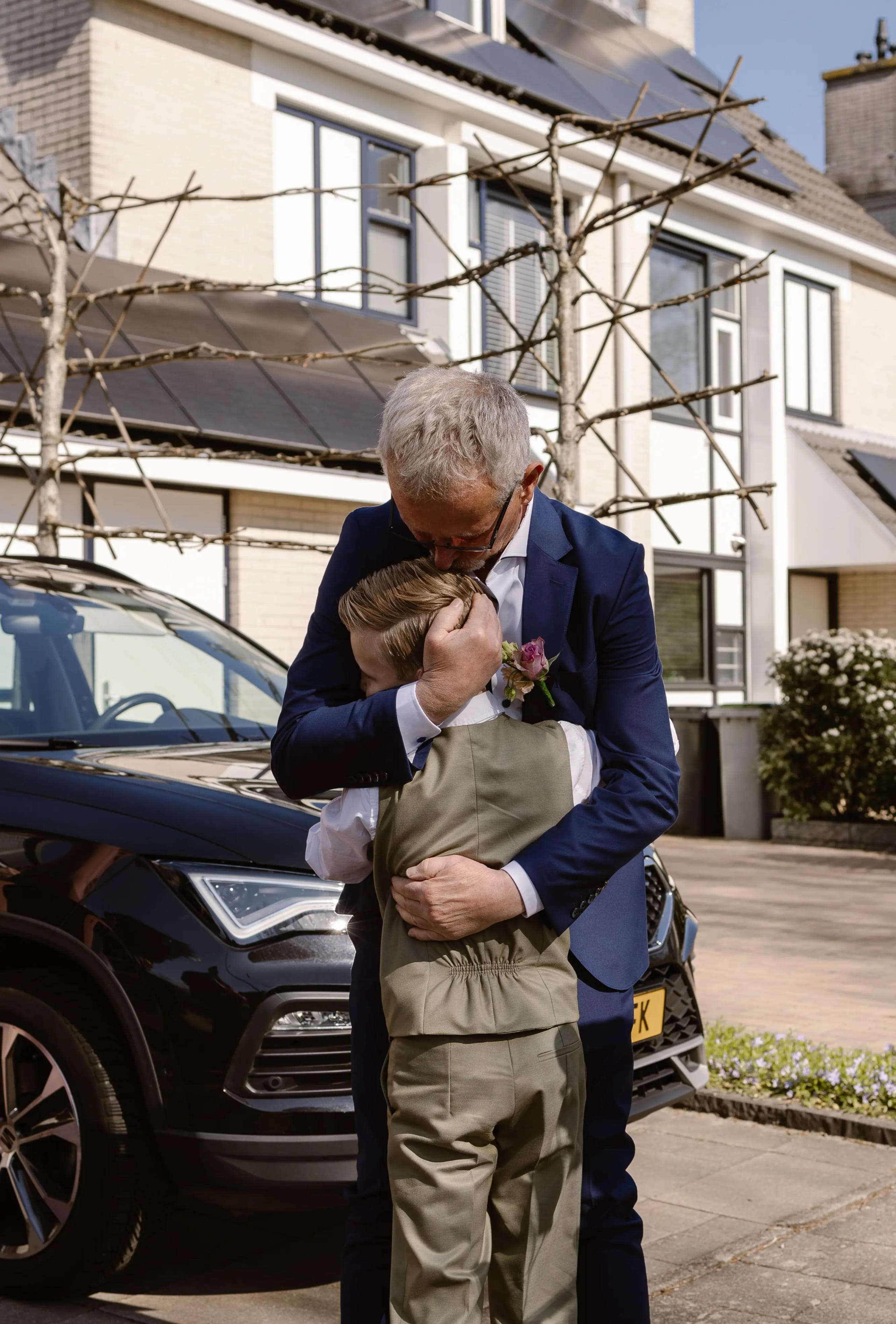 Bruidegom in pak geeft buiten een dikke knuffel aan het bruidskind naast een trouwauto
