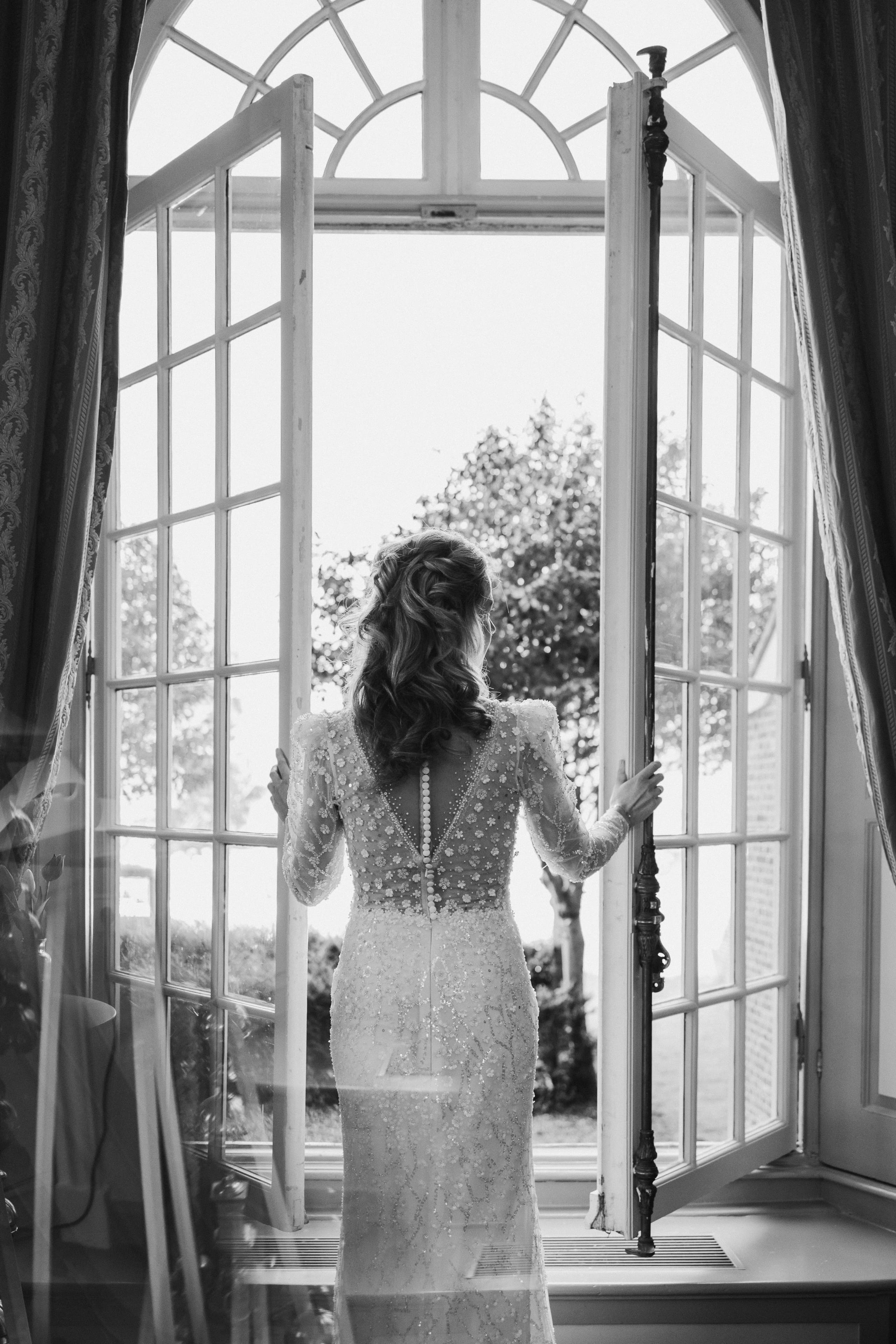 Zwart-wit foto van de bruid met open details op de rug die door de deuren naar buiten kijkt