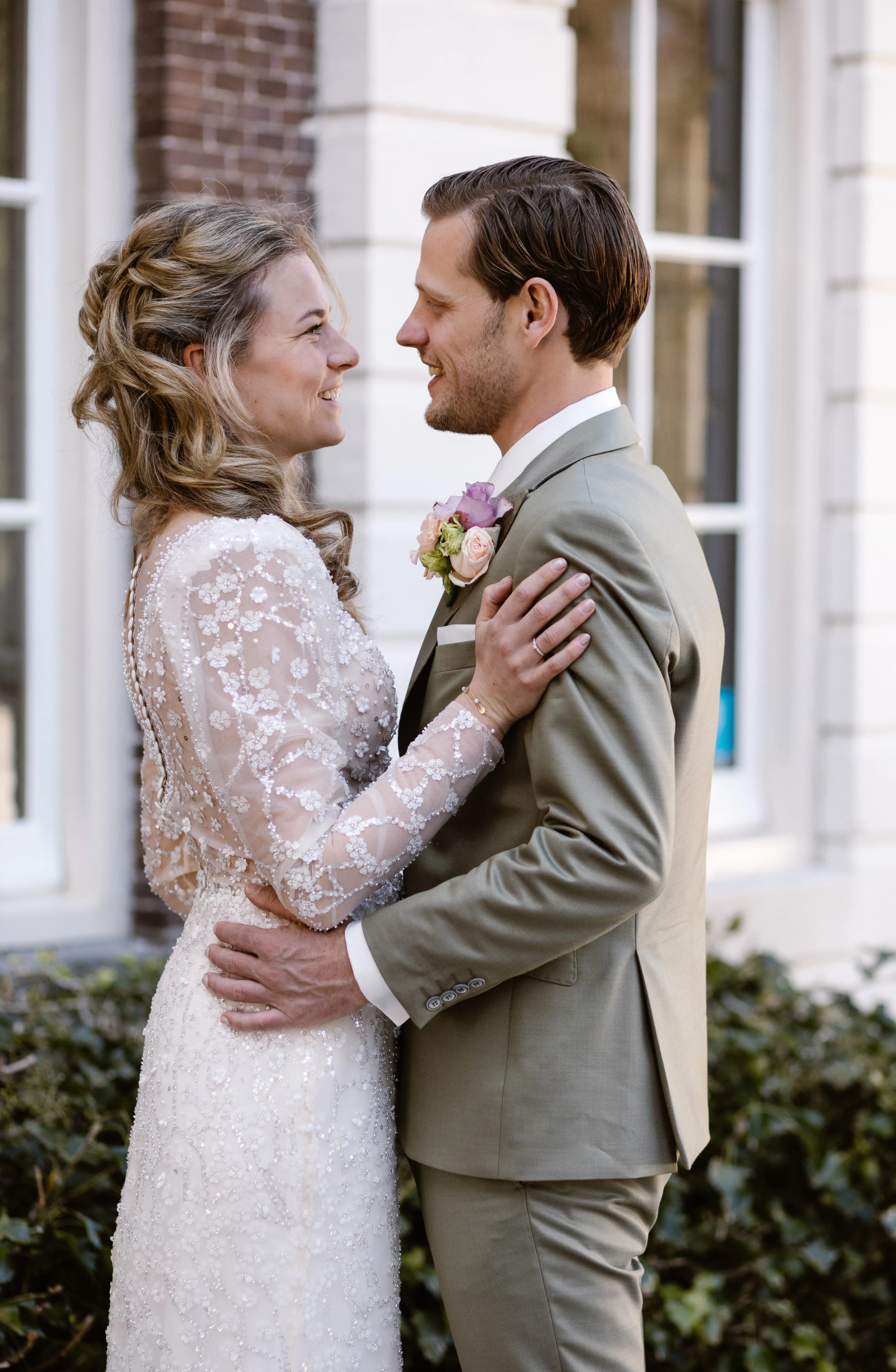 Gelukkig bruidspaar lacht elkaar stralend toe voor grote raampartijen binnen