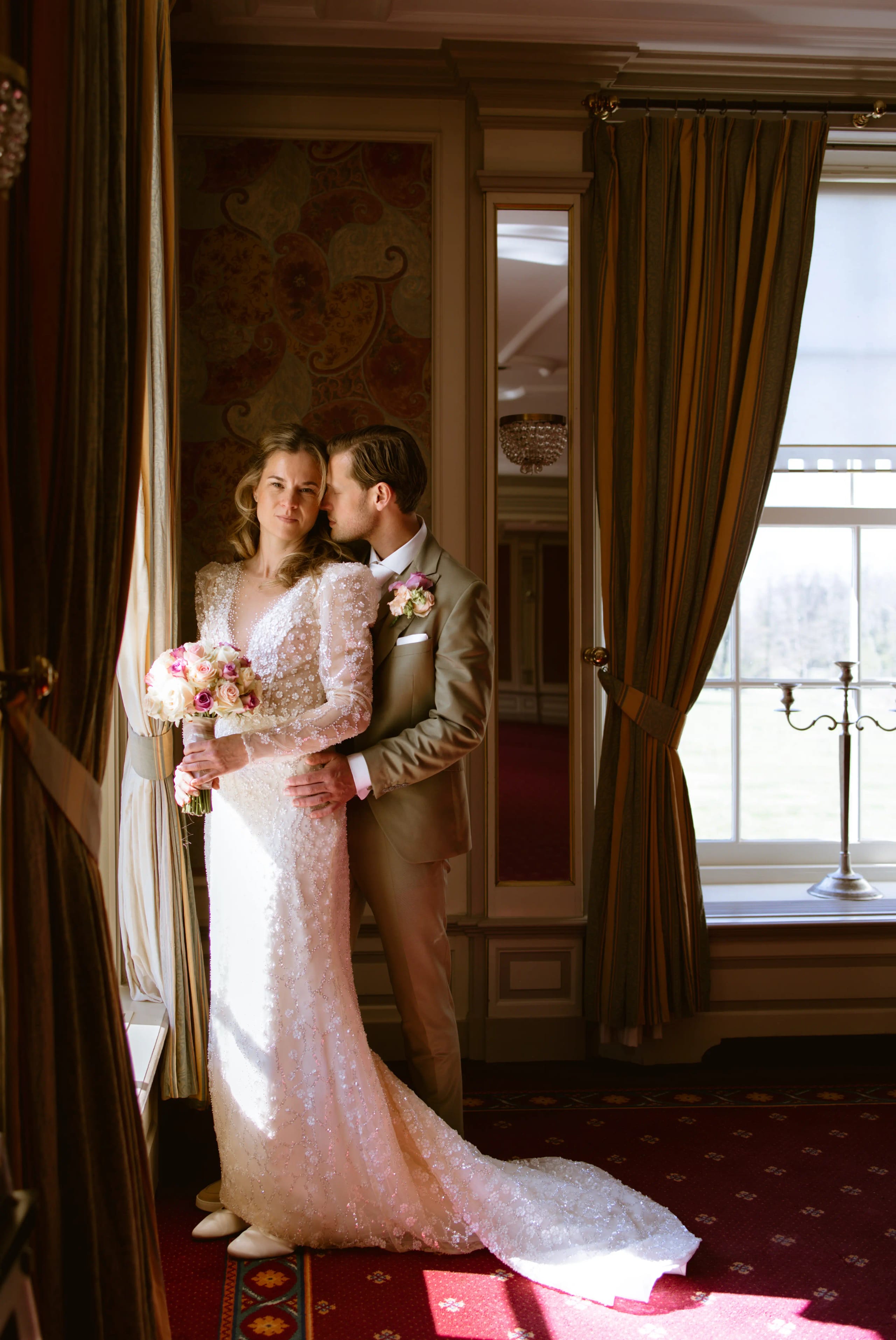 Zij-aan-zij pose van lachend bruidspaar hand in hand voor het raam van de kasteelzaal