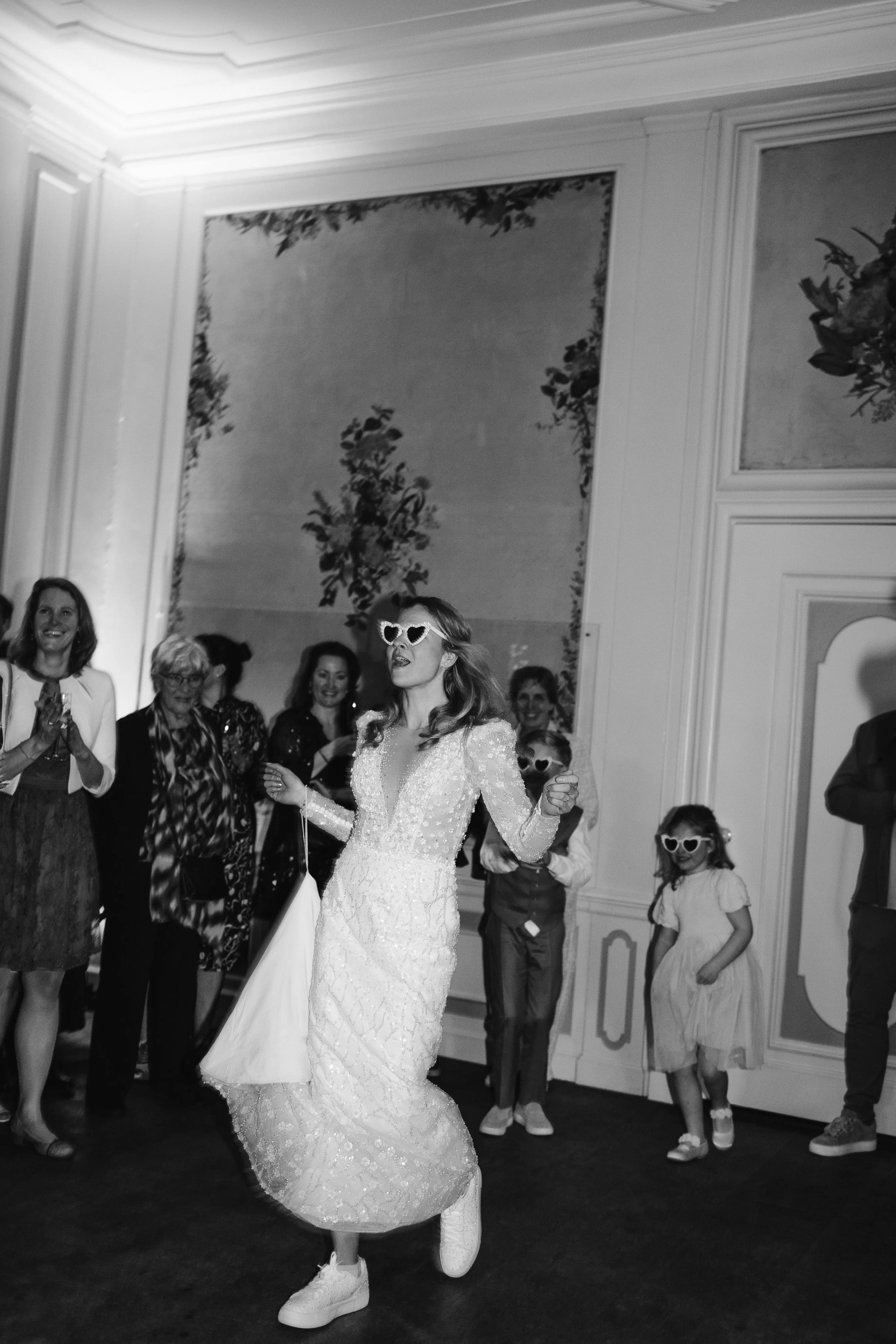 Zwart-wit actieshot van feestende bruid en kind die de dansvloer opkomen met hartjeszonnebrillen