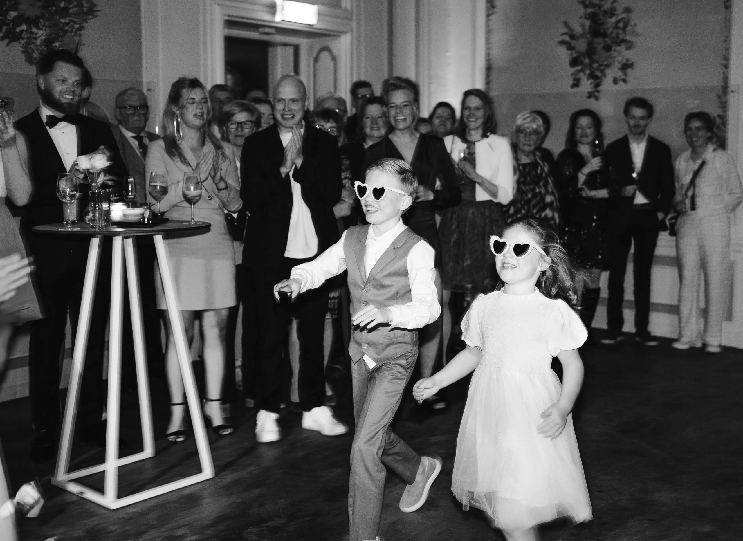 Zwart-wit actieshot van bruidegom en jongen die enthousiast dansend de zaal betreden in stijl