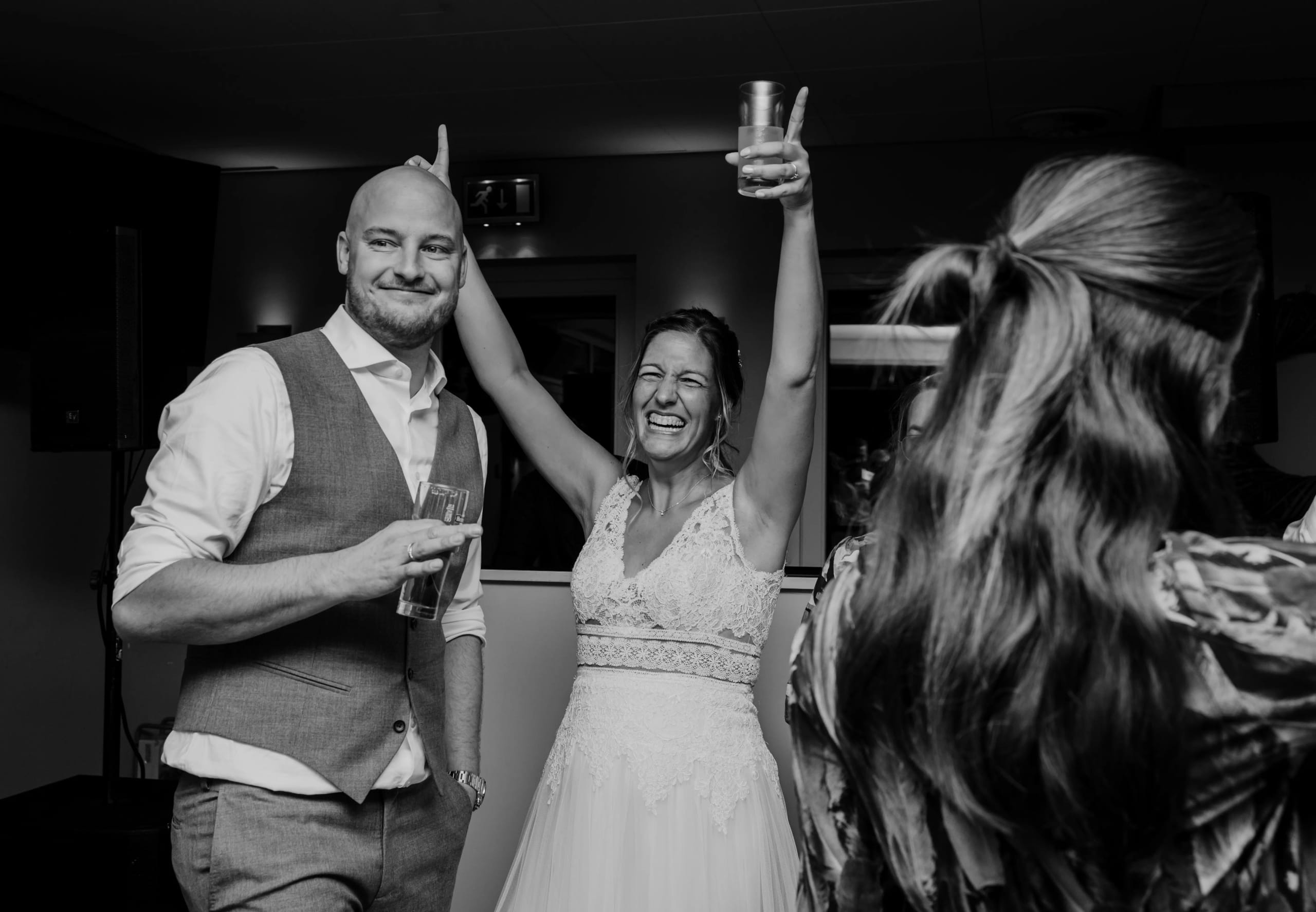 Bruidegom in grijs pak en bruid in kanten jurk lachen enthousiast met een drankje in de hand