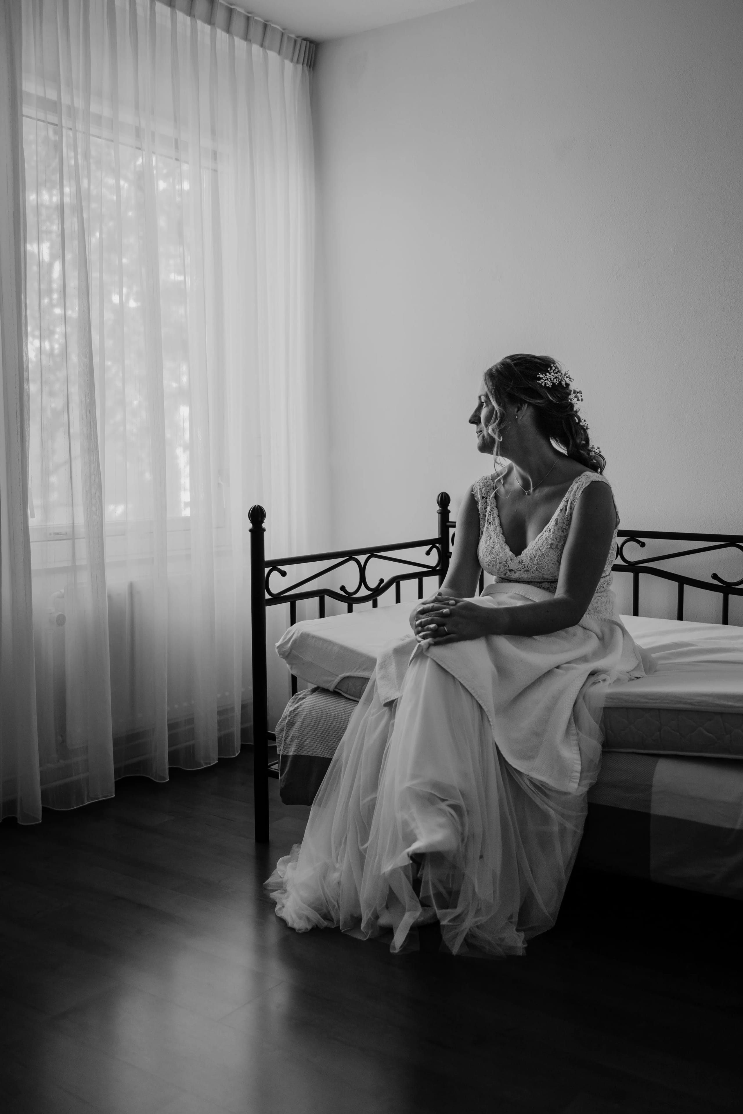 Zwart-wit portret van de bruid in haar trouwjurk zittend op de rand van een bed in een lichte kamer
