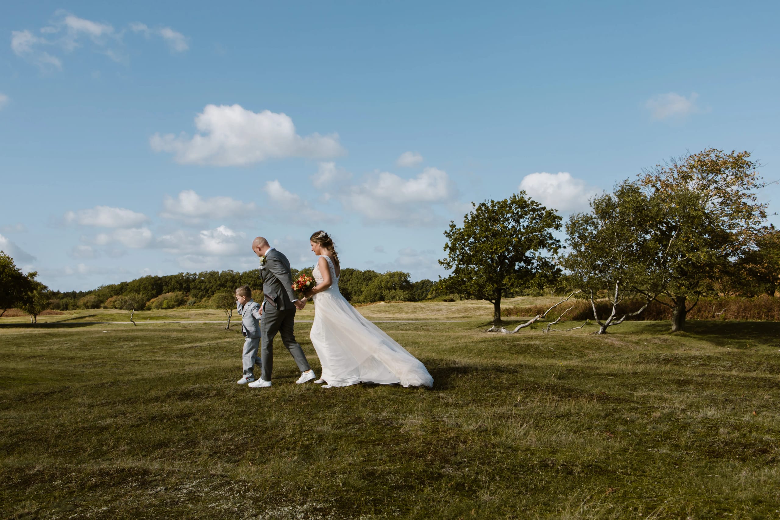 Het bruidspaar en een bruidsjonkertje wandelen hand in hand door een weids natuurgebied