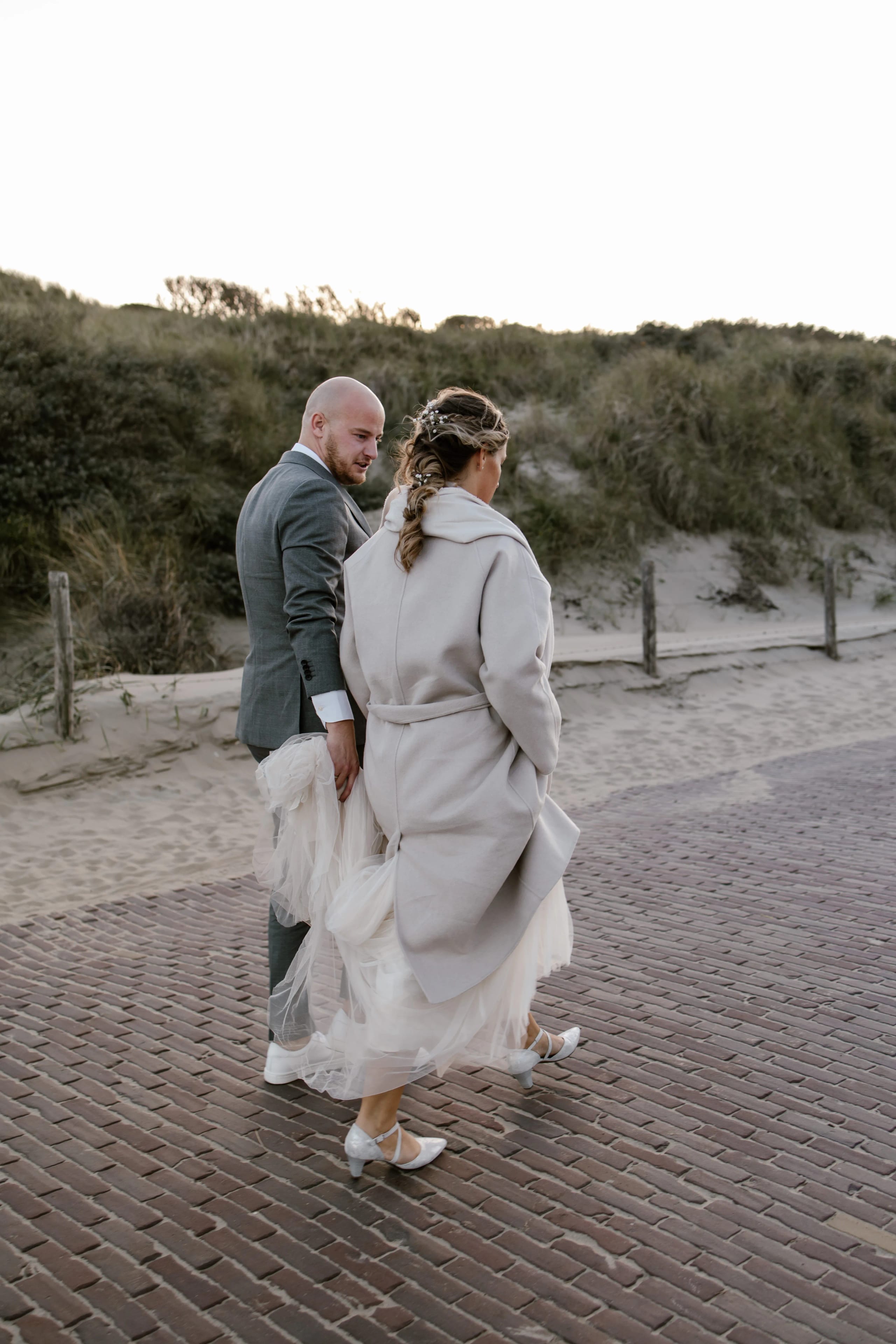 De bruid in haar trouwjurk met een lange mantel wandelt met de bruidegom door de duinen