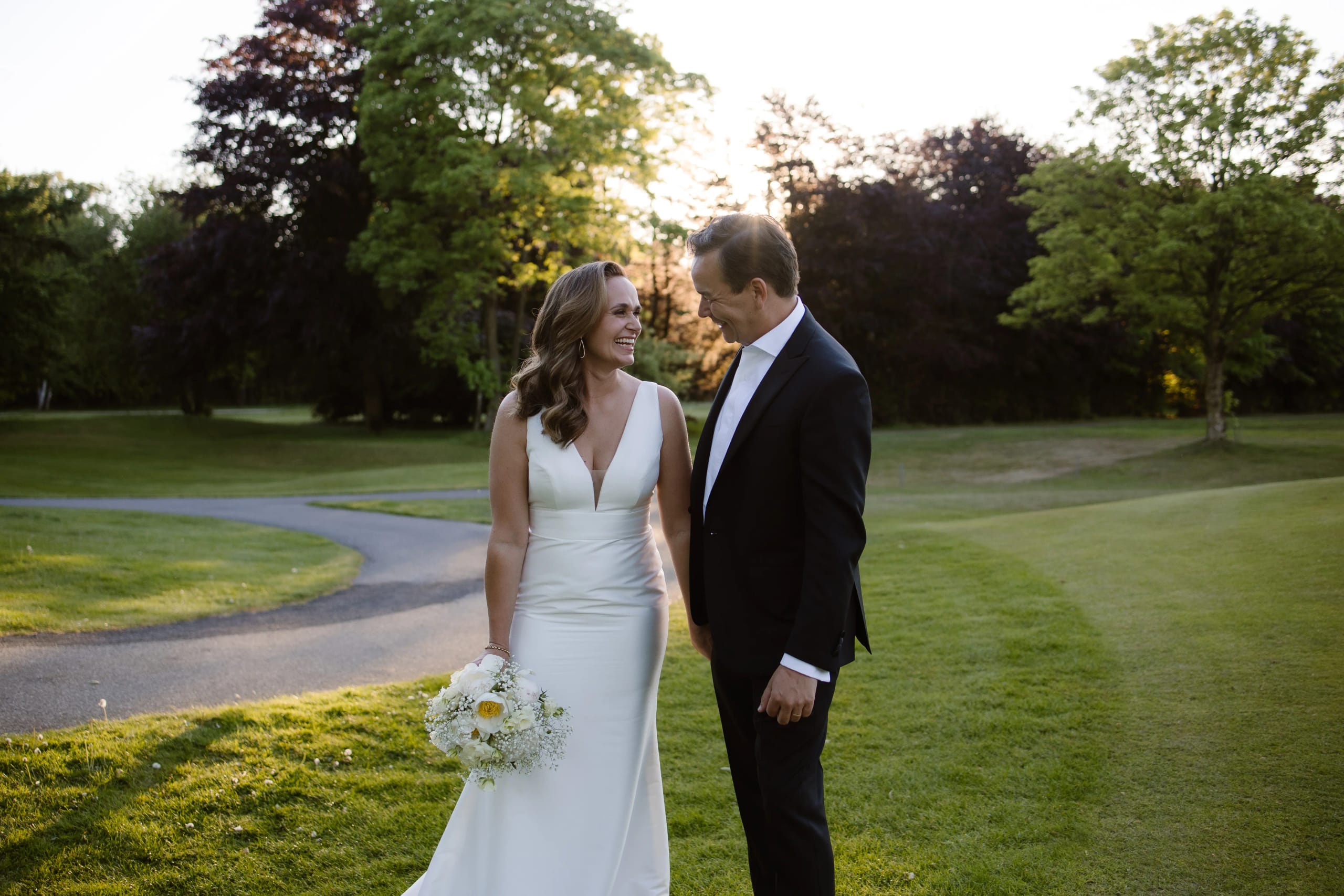 Bruid en bruidegom kijken elkaar lachend aan terwijl ze hand in hand door de tuin lopen