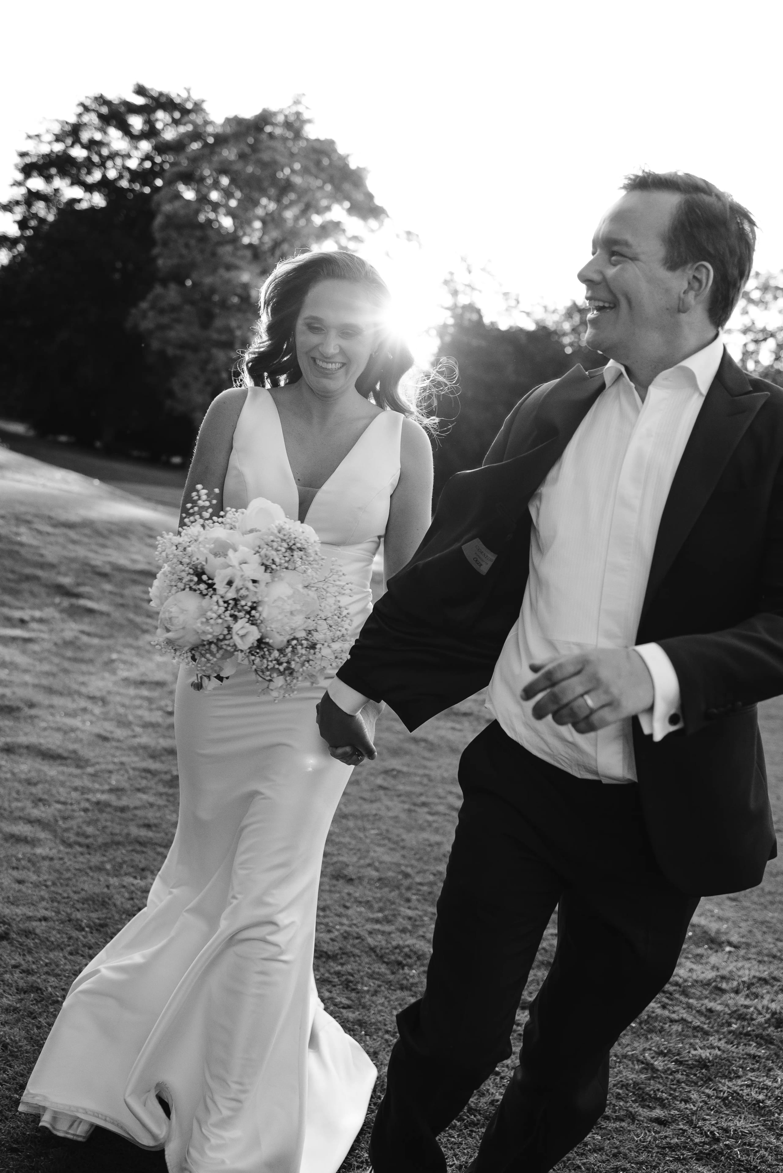 Lachend bruidspaar loopt hand in hand in de zon, zwart-wit fotografie