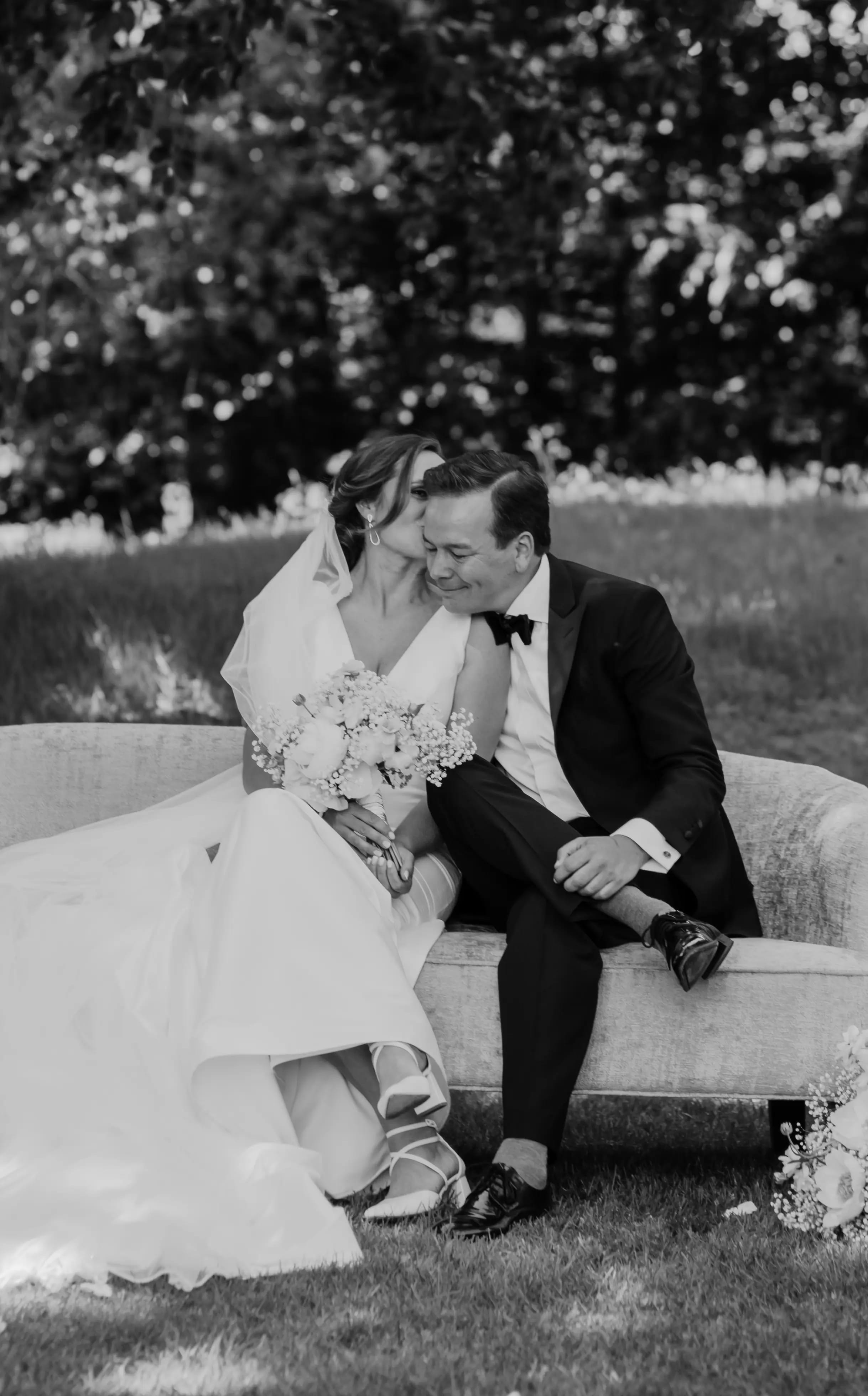 Zwart-wit foto van het bruidspaar dat elkaar een kus geeft zittend op een stenen bankje
