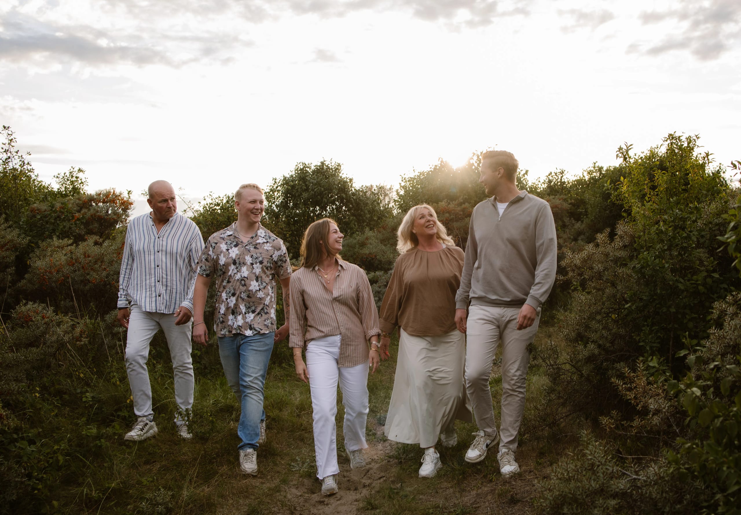 Een groep van vijf volwassenen wandelt samen op een schilderachtig pad omringd door groen, met een zonsondergang op de achtergrond. De mensen dragen casual kleding en lachen terwijl ze met elkaar in gesprek zijn, wat een gevoel van saamhorigheid en vreugde uitstraalt.