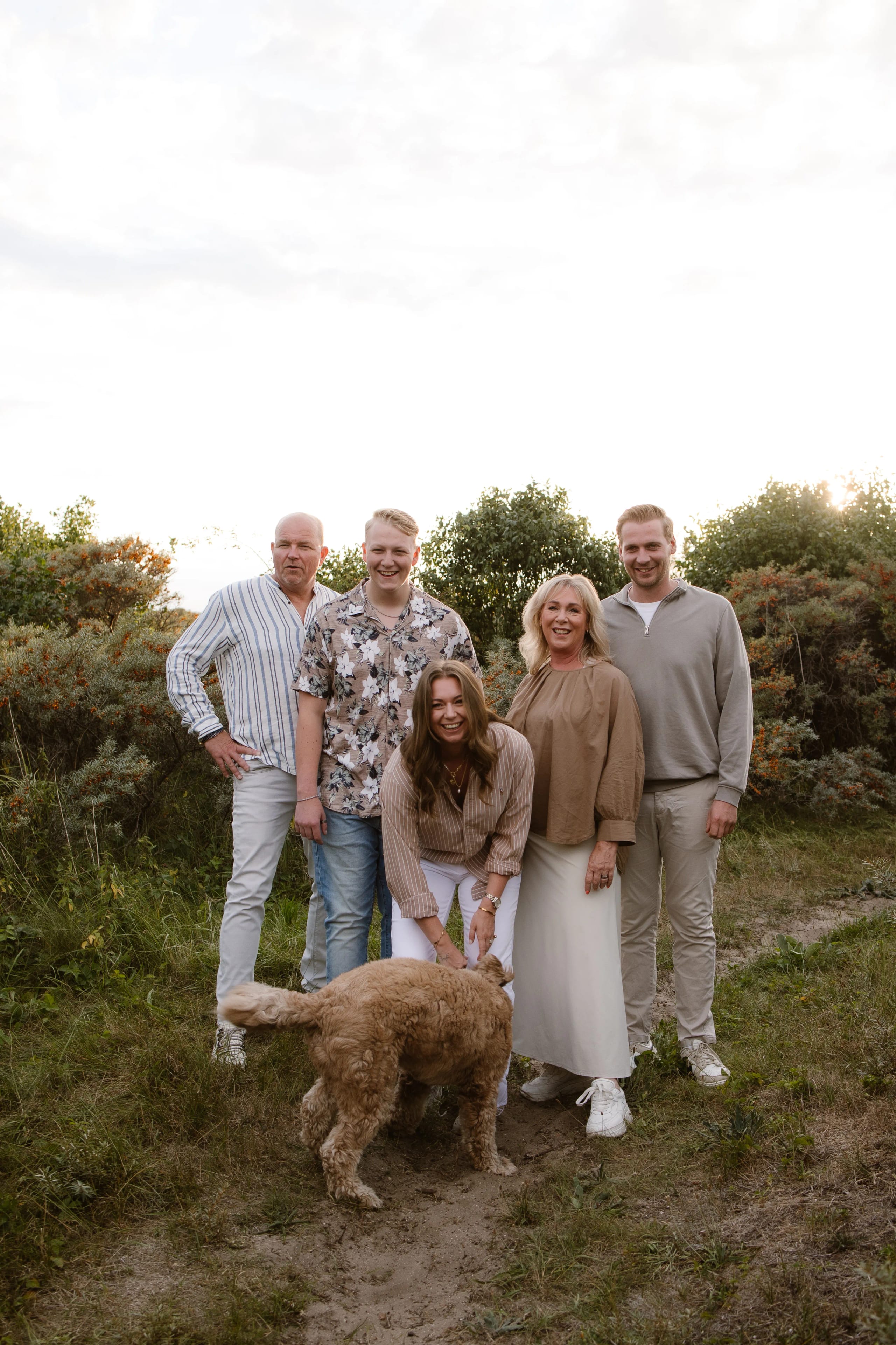 Een vrolijke gezinssamenstelling met vijf personen, waaronder een vrouw die met een hond speelt, staan samen in een groene omgeving met struiken op de achtergrond, onder een bewolkte lucht.