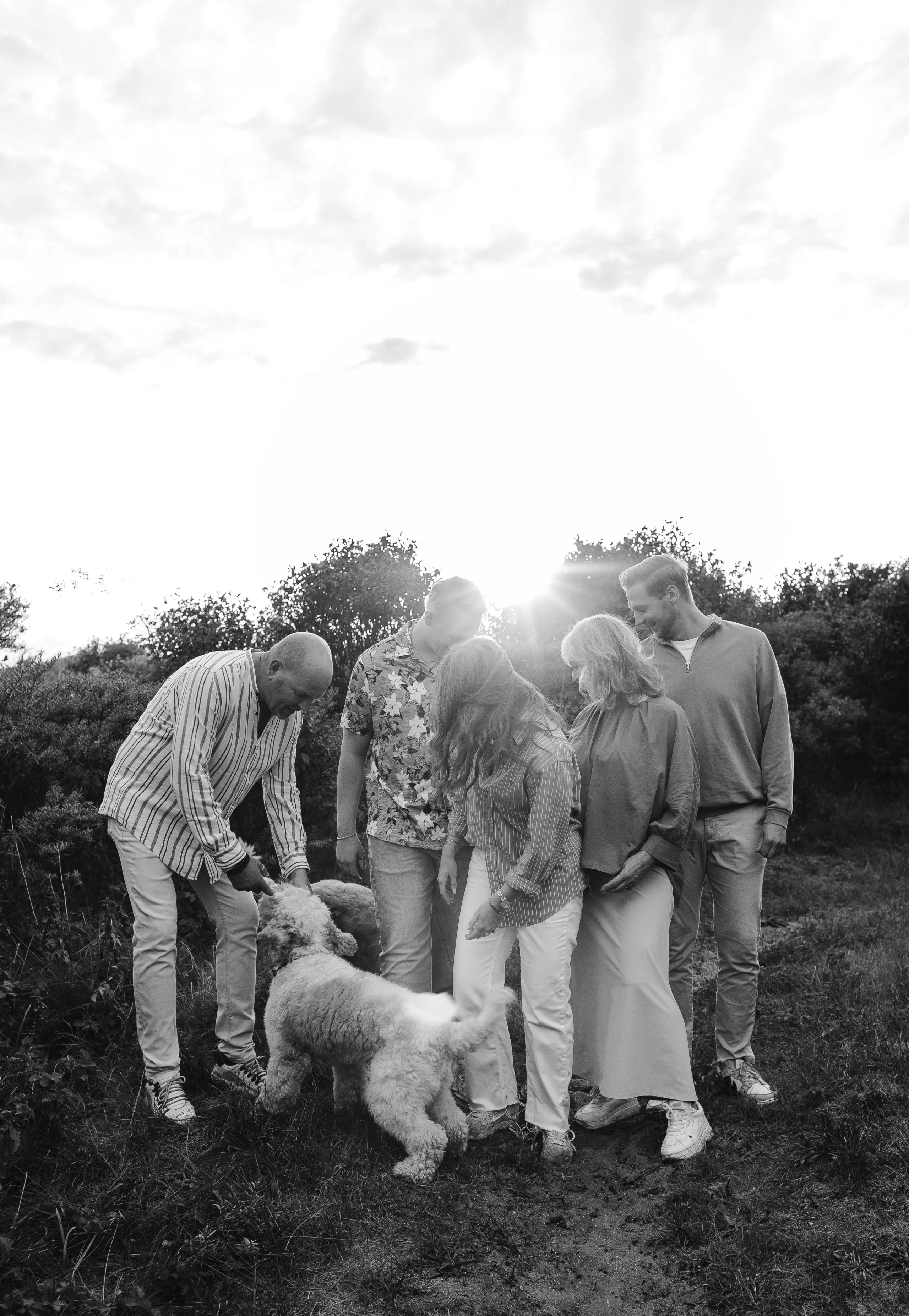 Een groep van vijf mensen staat samen in een groene omgeving, terwijl een hond tussen hen doorloopt en hun aandacht trekt; de zon schittert op de achtergrond, waardoor een warme sfeer ontstaat.