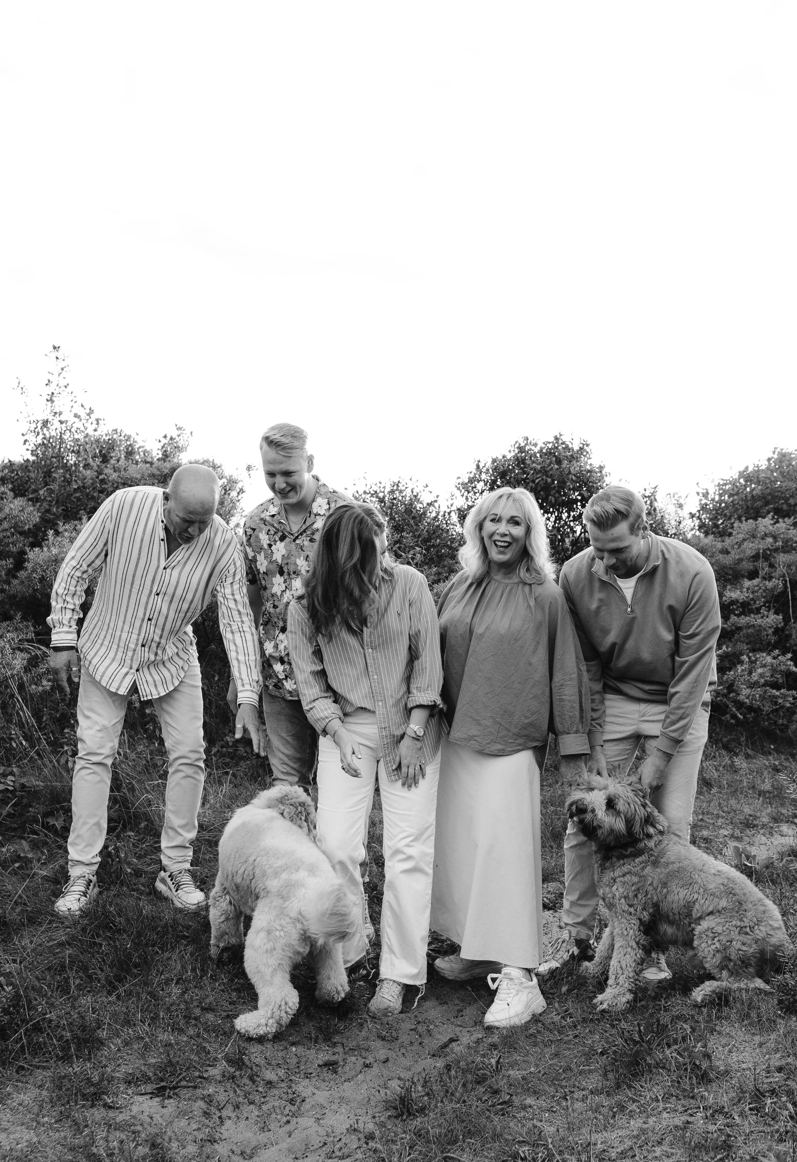 Een groep van vijf mensen, gekleed in informele mode, staat buiten op een grasveld en lacht terwijl ze naar twee honden kijken. De sfeer is vrolijk en informeel, met groenachtige bomen op de achtergrond.