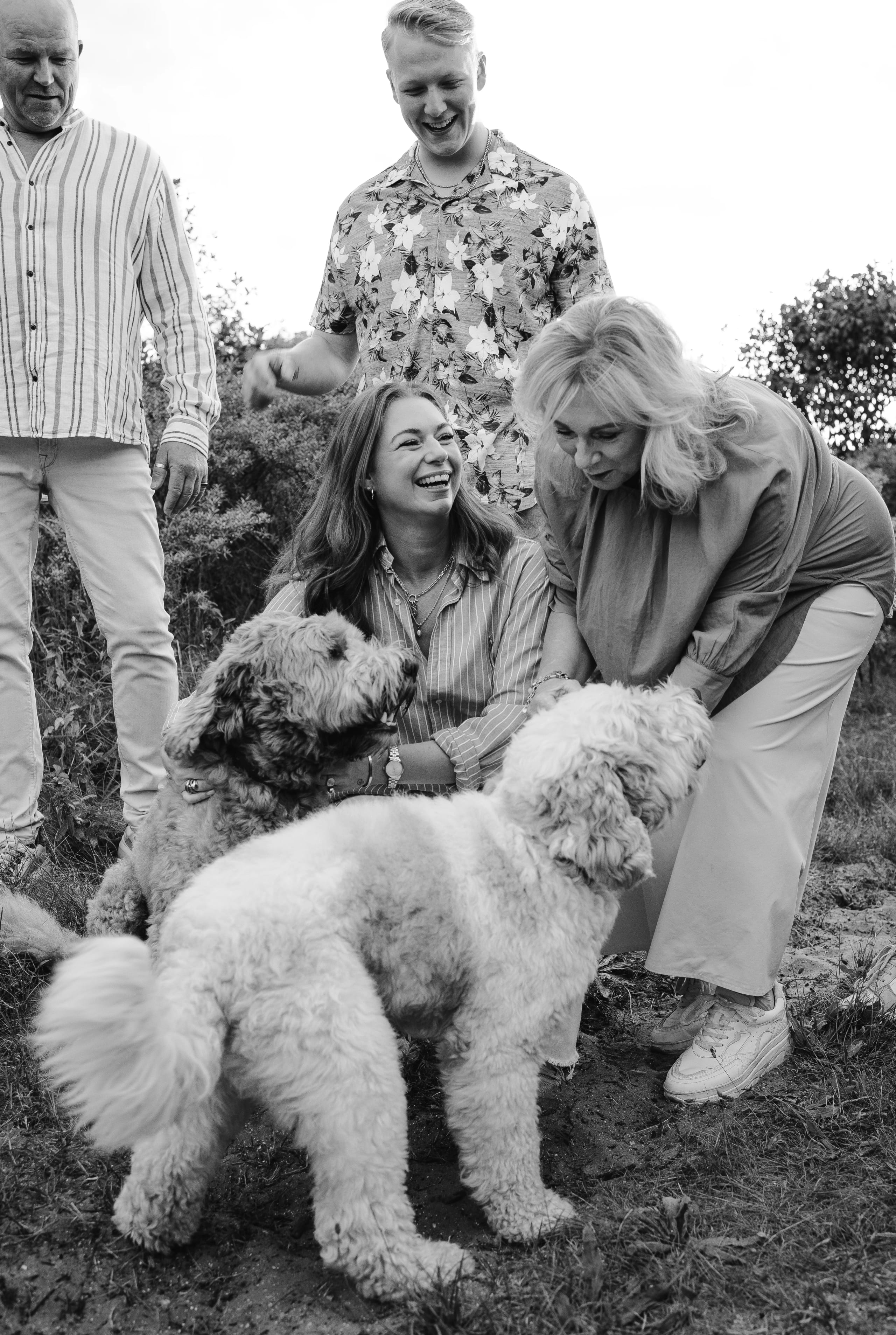 Vijf mensen genieten van een vrolijke interactie met twee honden in een groene buitenomgeving, waarbij ze lachen en elkaar vriendelijk berijden.