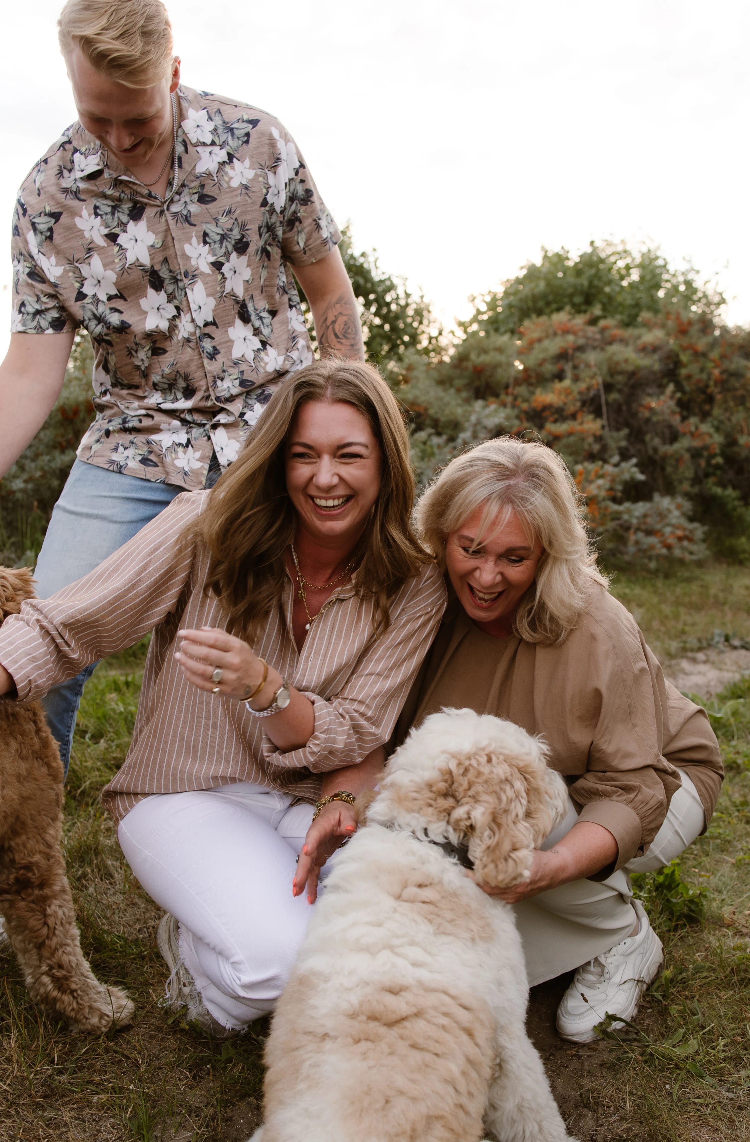 Vijf mensen genieten van een gezellige bijeenkomst in de natuur, lachen en spelen met twee honden, terwijl ze in een groene omgeving staan. De drie vrouwen, gekleed in casual mode, stralen blijheid uit, terwijl een man zich op de achtergrond bij hen voegt.