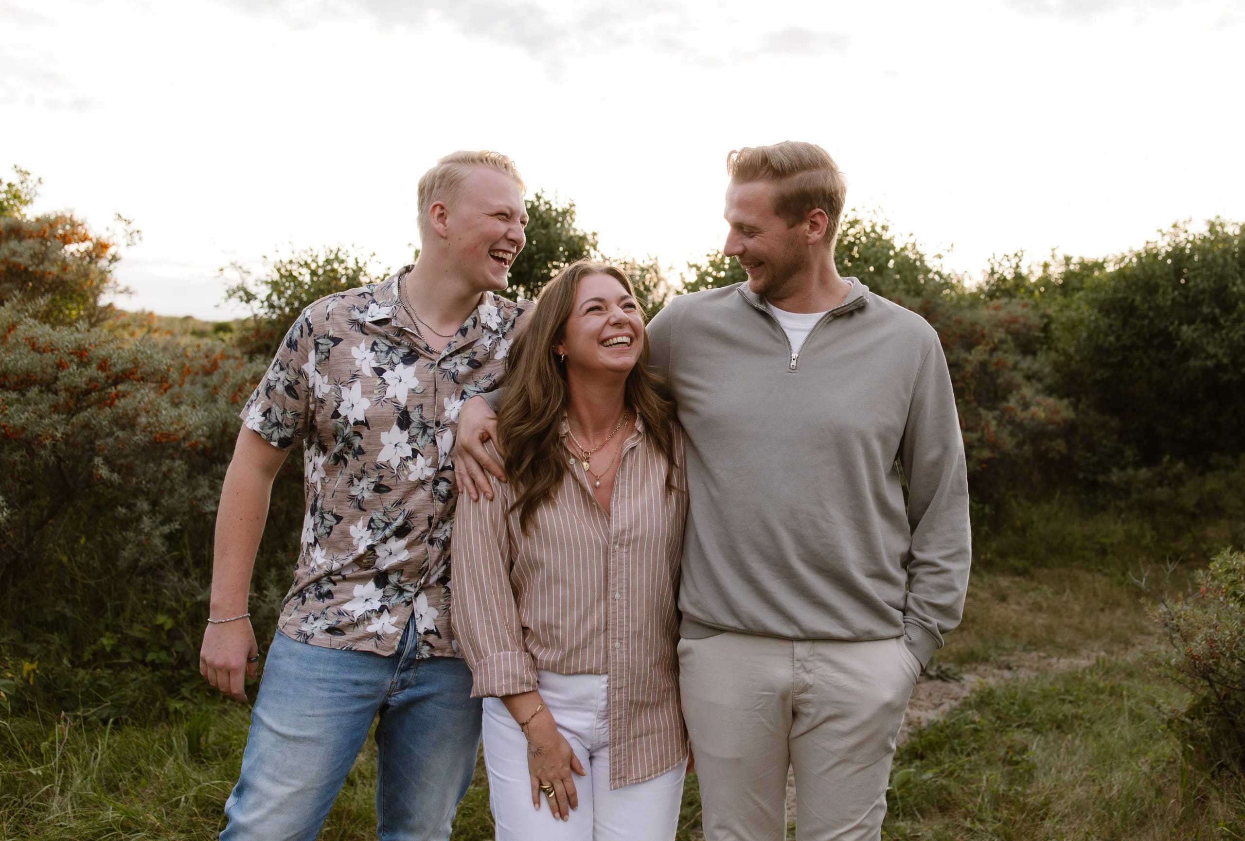 Drie mensen lachen en genieten van elkaar in een groene, natuurlijke omgeving tijdens een zonsondergang, met een vrouw in een gestreepte blouse en twee mannen in casual kleding die elkaar knuffelen en kletsen.