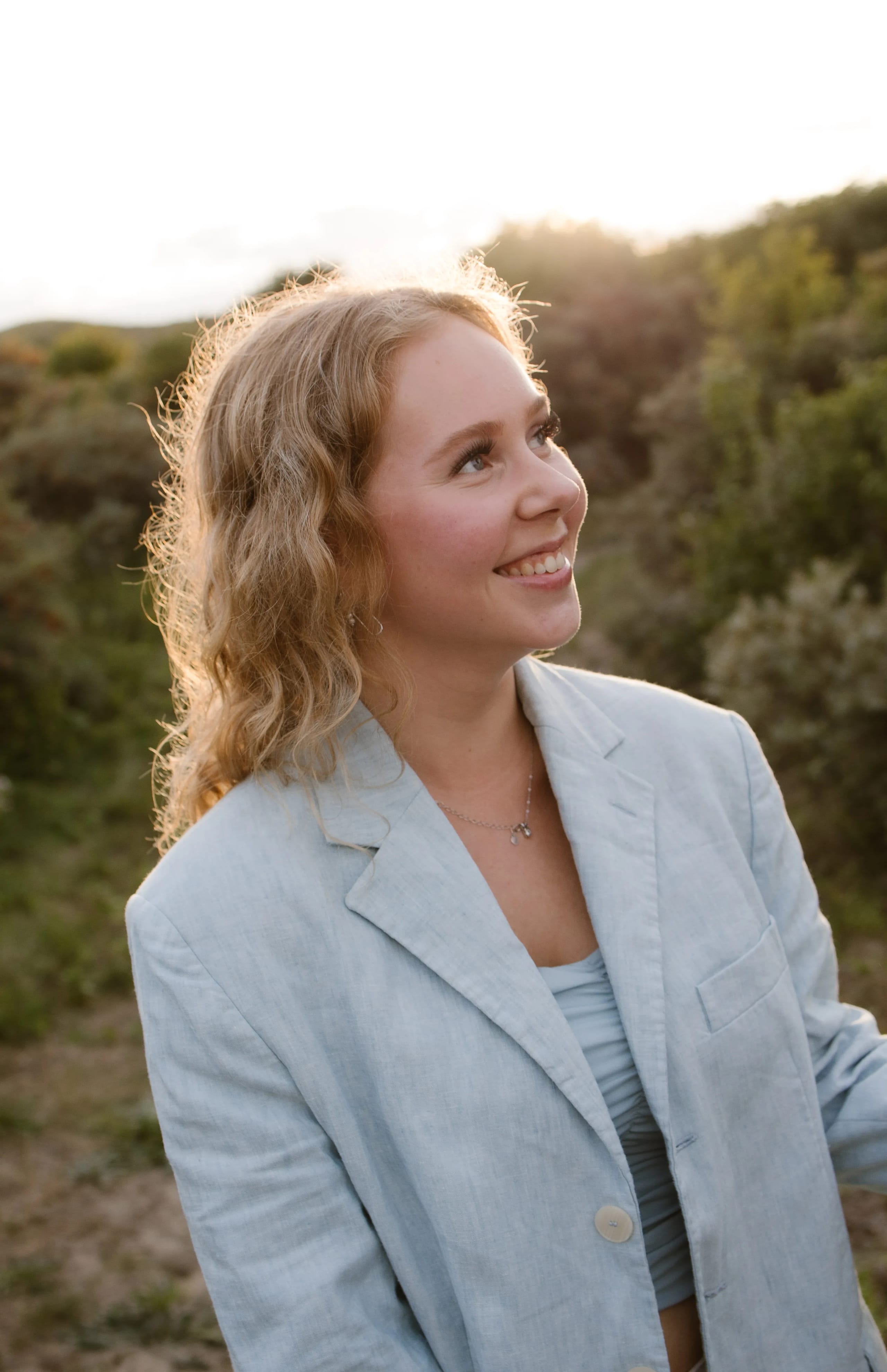 Een jonge vrouw met krullend haar, gekleed in een lichtblauw lakenjack en een top, glimlacht terwijl ze naar de horizon kijkt in een groene omgeving bij zonsondergang.