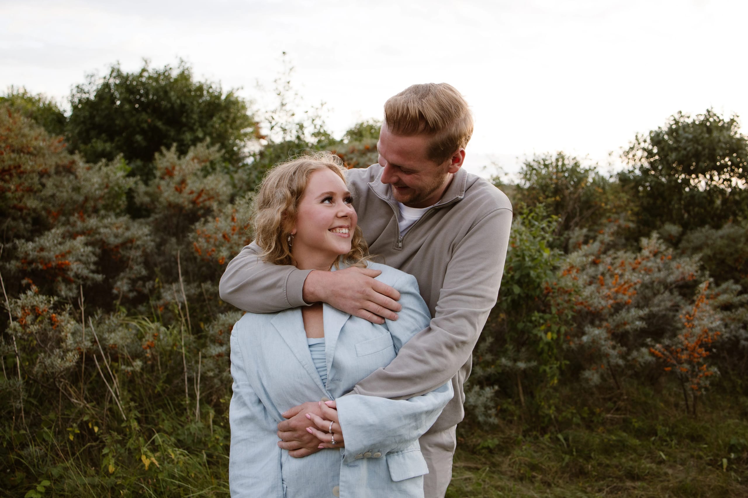 Een stel omhelst elkaar vrolijk in een natuurlijke omgeving met groene struiken en een zachte zonsondergang op de achtergrond, waarbij ze elkaar liefdevol aankijken.