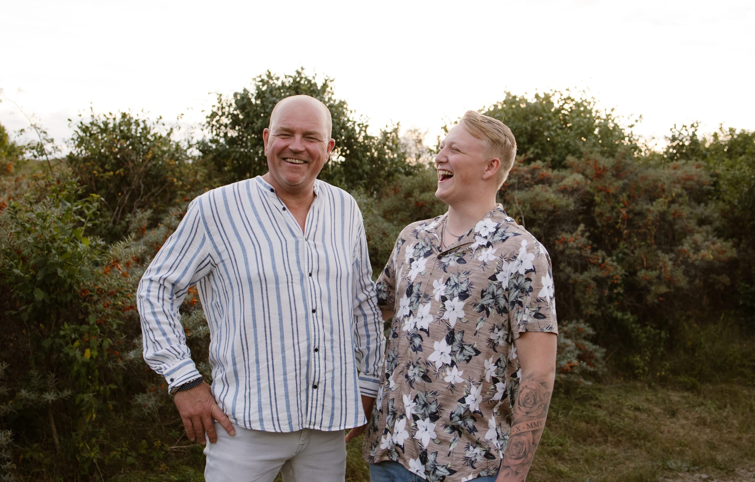 Twee mannelijke familieleden lachen samen in een groene omgeving, omringd door struiken en bomen. De man links draagt een gestreepte overhemd, terwijl de man rechts een shirt met bloemenprint aan heeft, wat een gezellige en ontspannen sfeer uitstraalt.