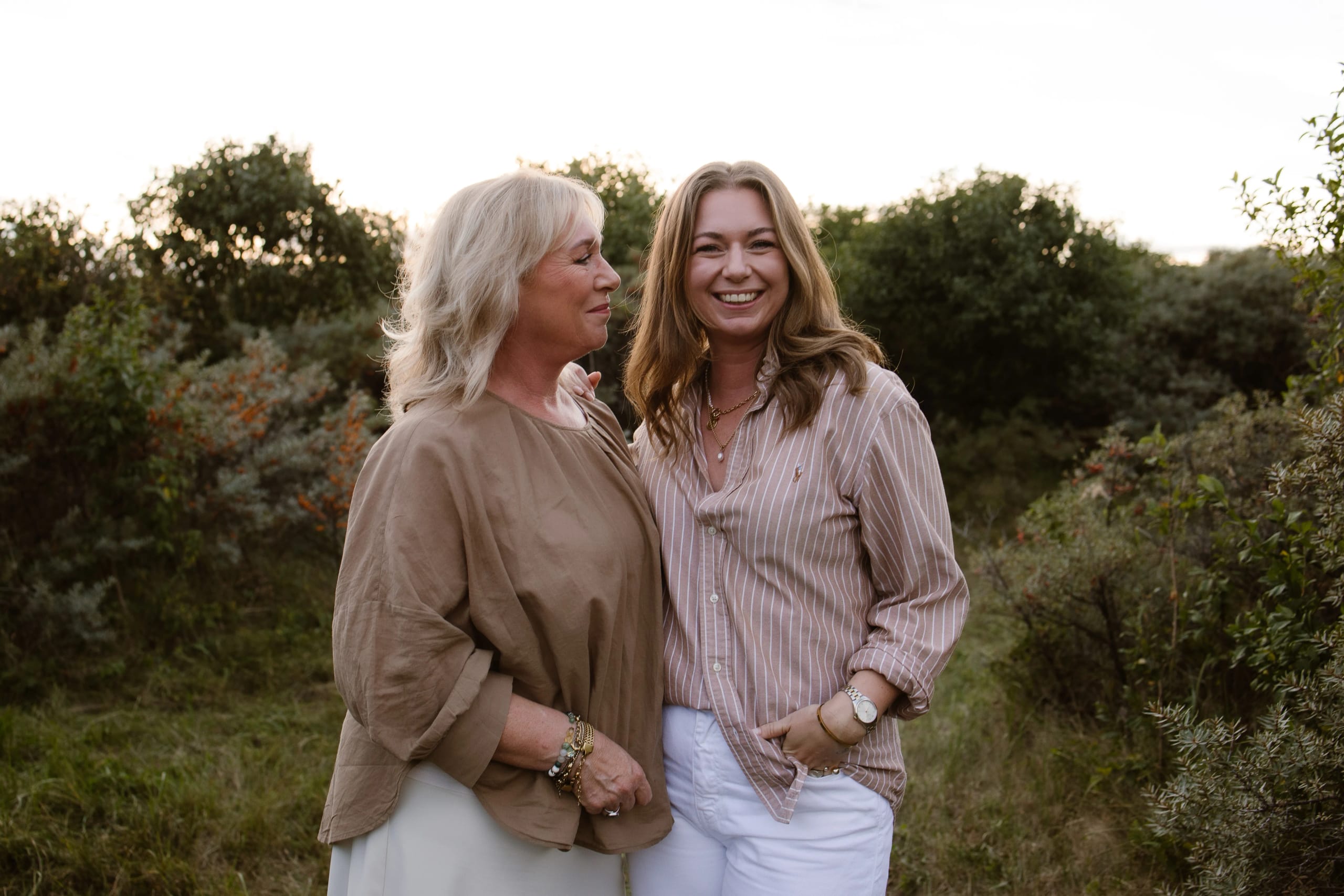 Twee vrouwen staan dicht bij elkaar in een groene omgeving, lachend en in gesprek terwijl de zon ondergaat. De vrouw links draagt een beige top, terwijl de vrouw rechts gekleed is in een gestreepte blouse.